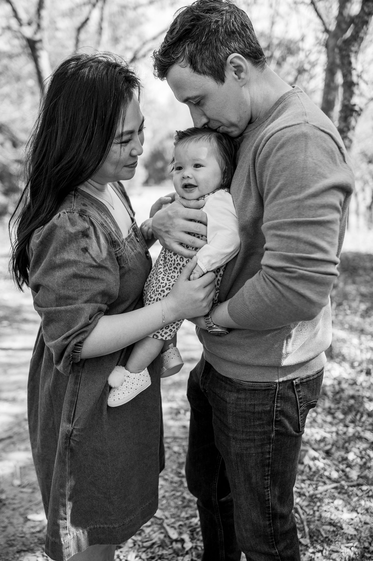 Black and white photo of a couple lovingly holding their smiling baby outdoors in a park setting.
