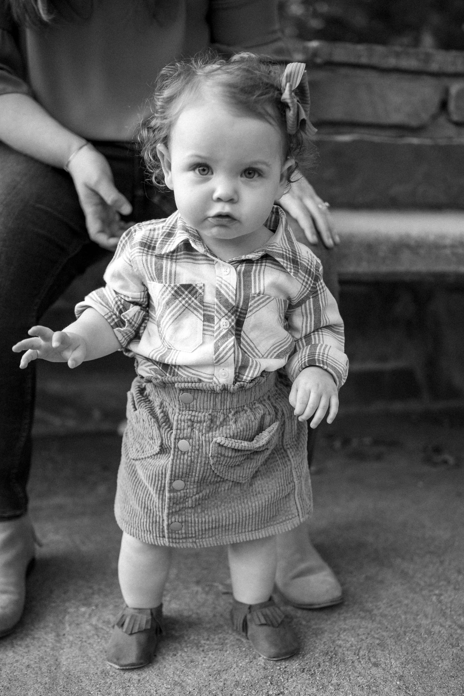 Black and white portrait of toddler in plaid shirt and corduroy skirt standing on steps.