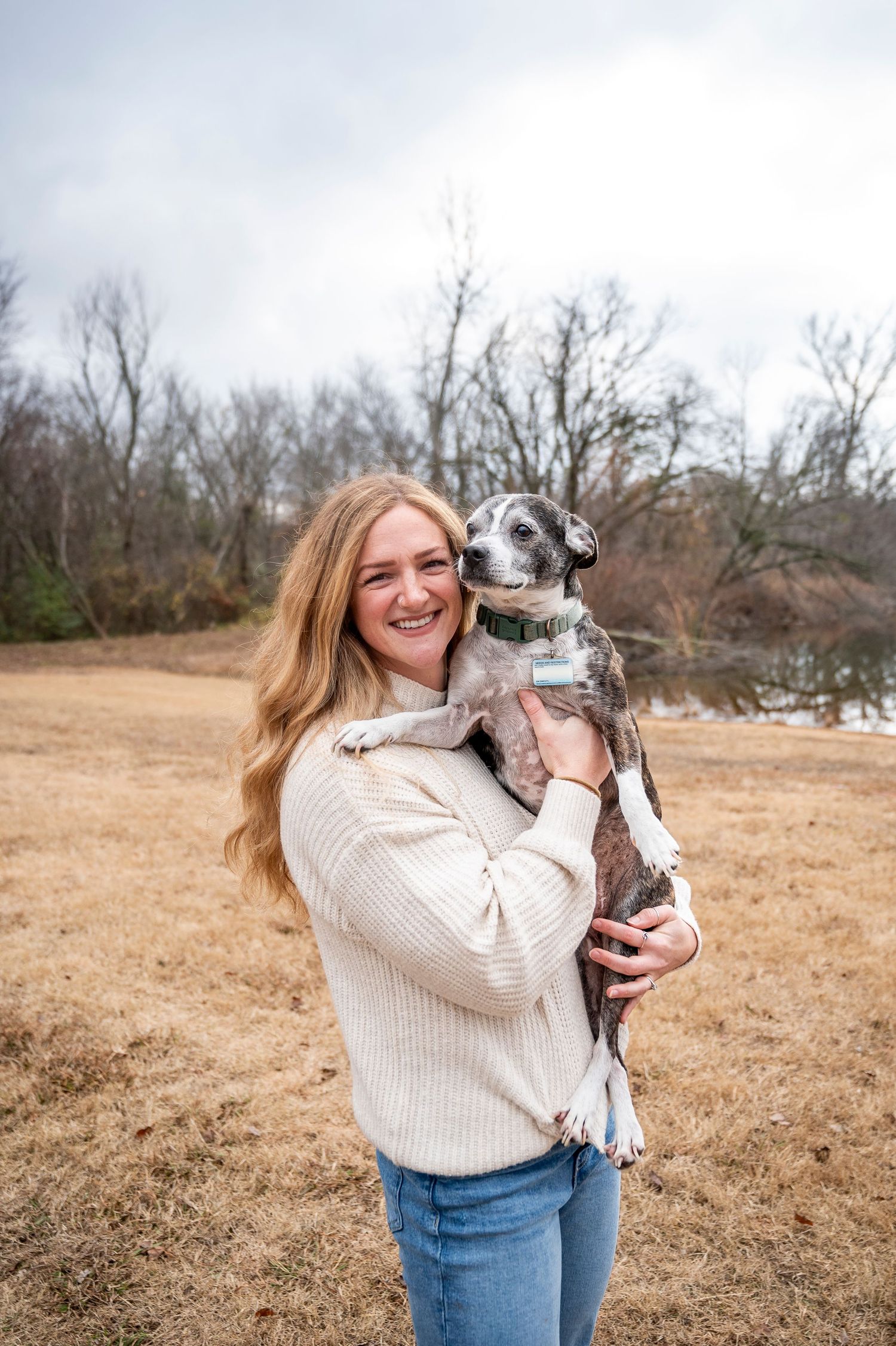 Person in cream sweater holding grey dog outdoors by lake.
