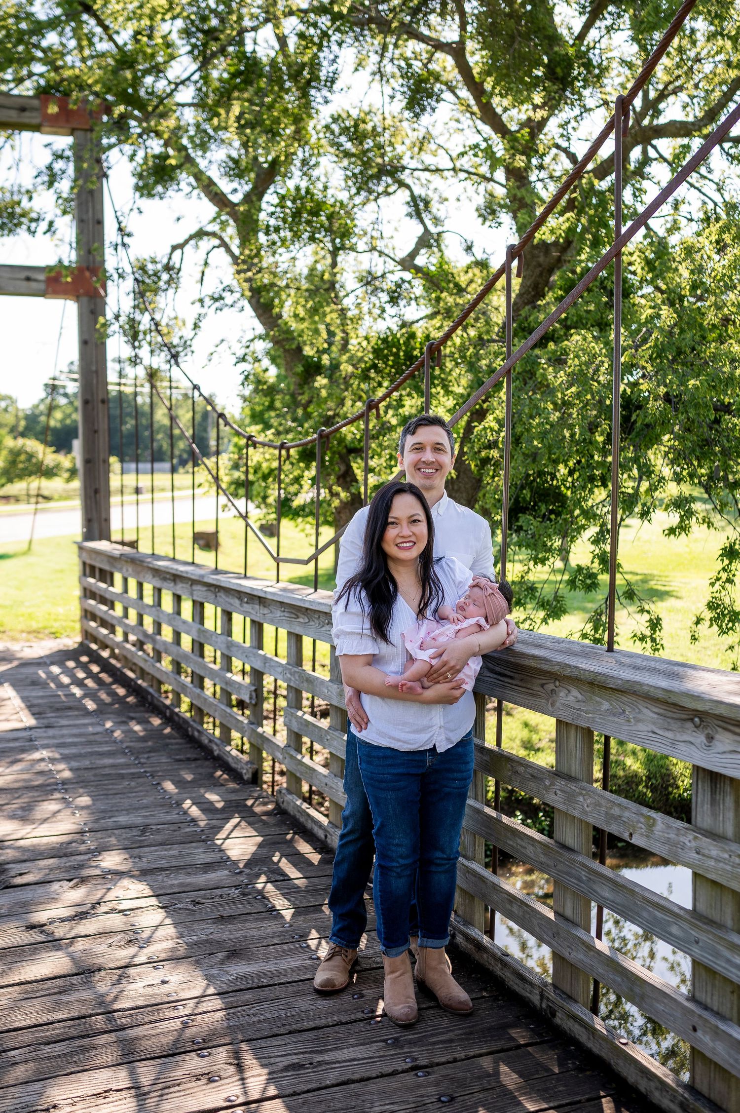 A couple stands together on a wooden bridge in a sunny outdoor setting surrounded by green trees.