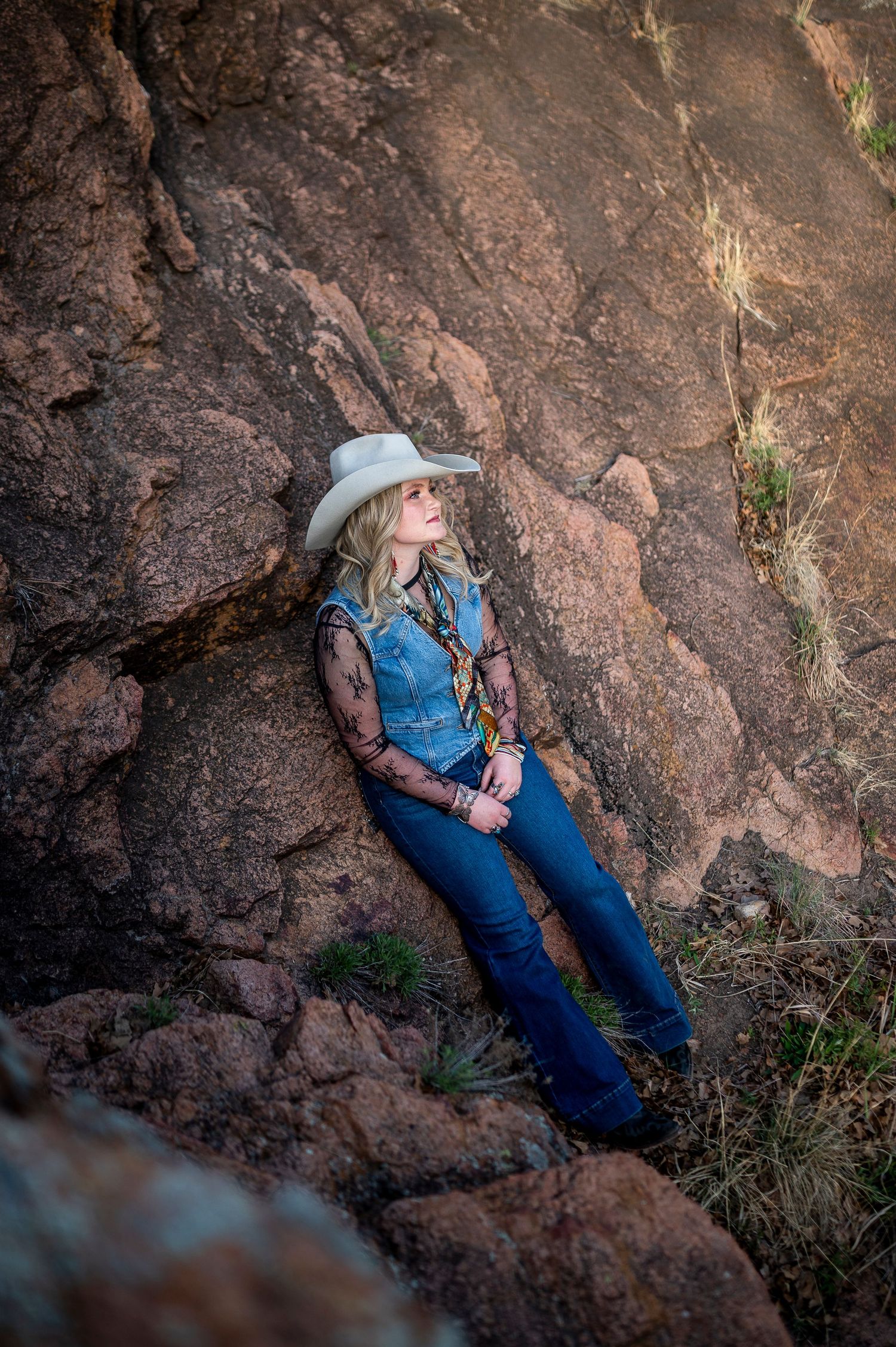 Blonde woman in white cowboy hat and denim vest leans against red rock cliff face looking upward outdoors.