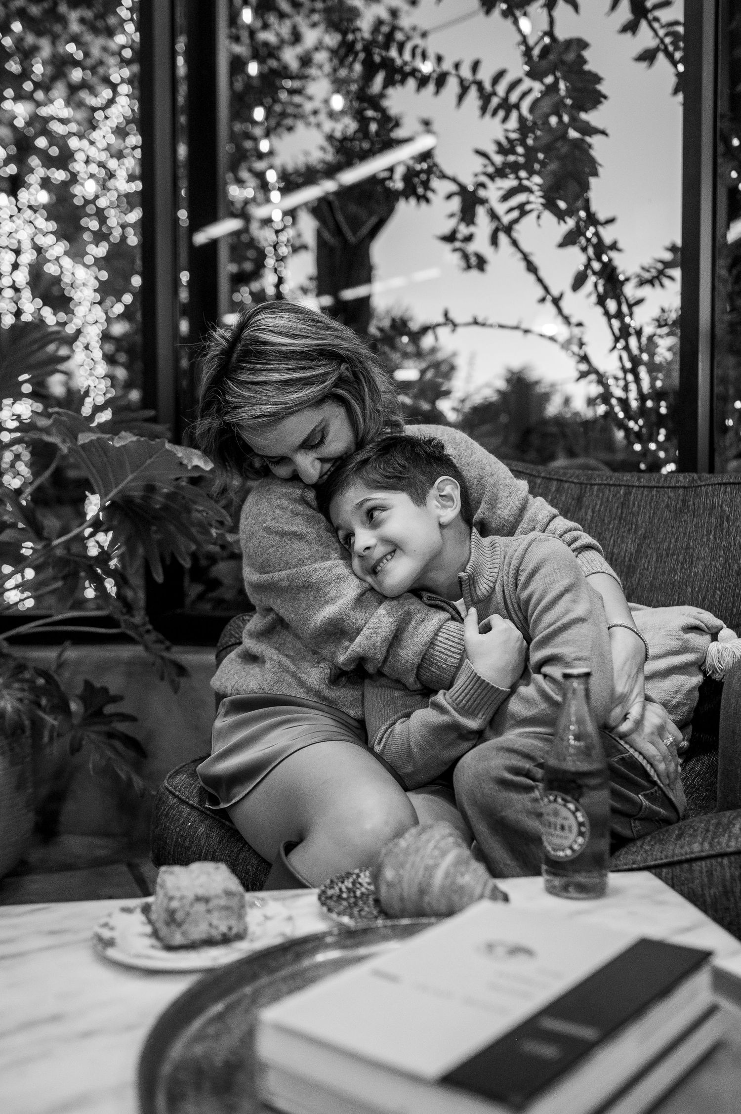 Woman hugging smiling boy on outdoor sofa with bokeh lights in black and white photo.