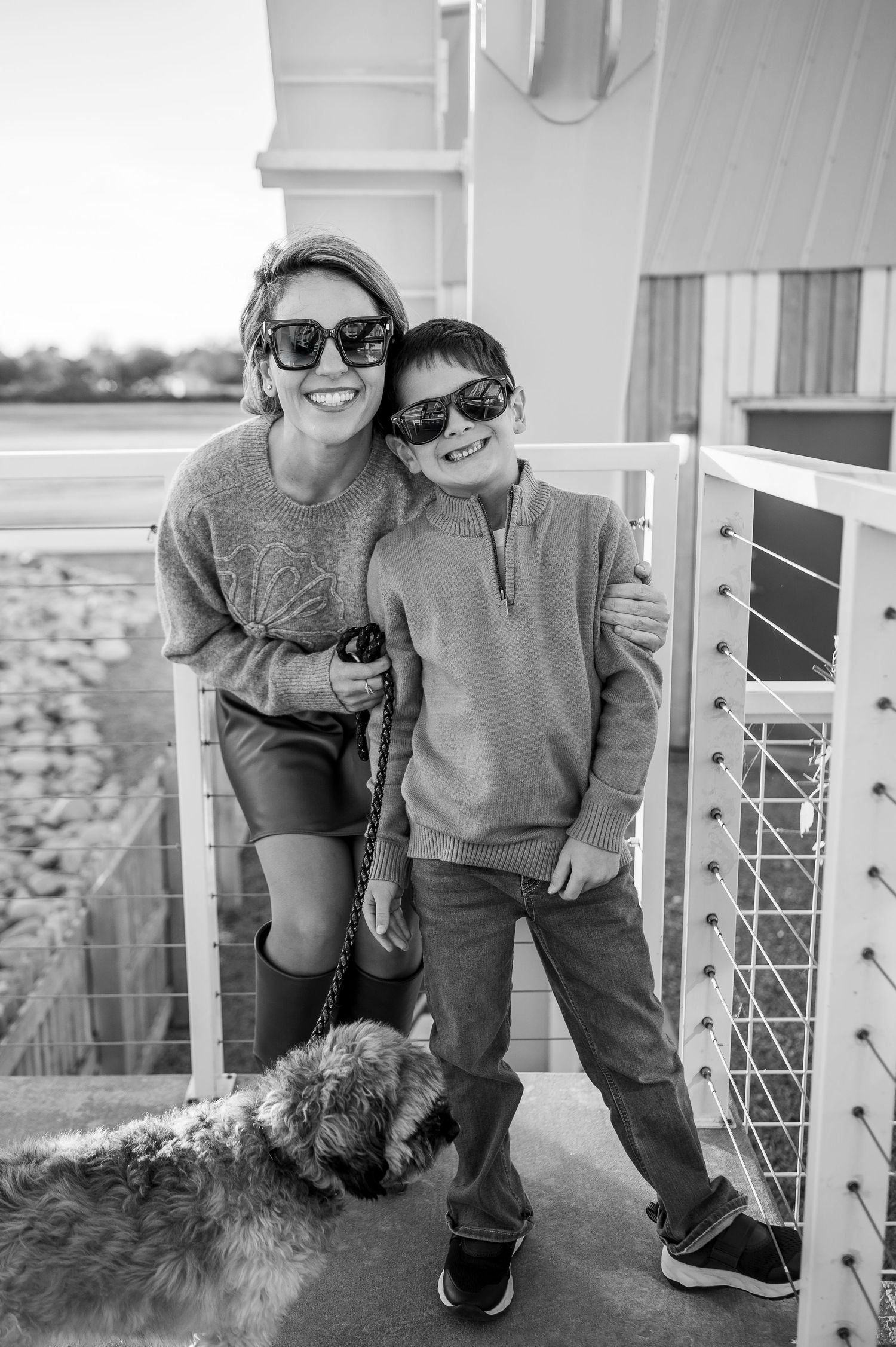 Woman and boy in sunglasses smile on a balcony with a dog in a black and white photo.