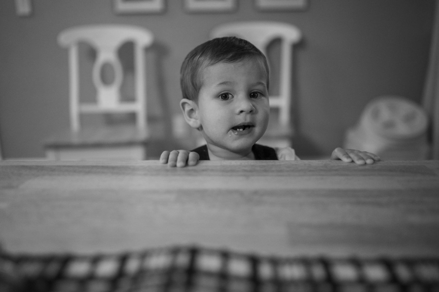 A young child peeks over a table edge in a black and white portrait.