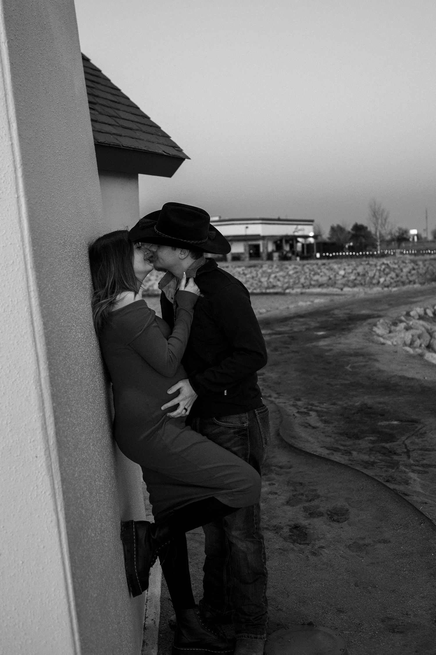 A romantic black and white photo of a couple in western wear embracing against a wall.