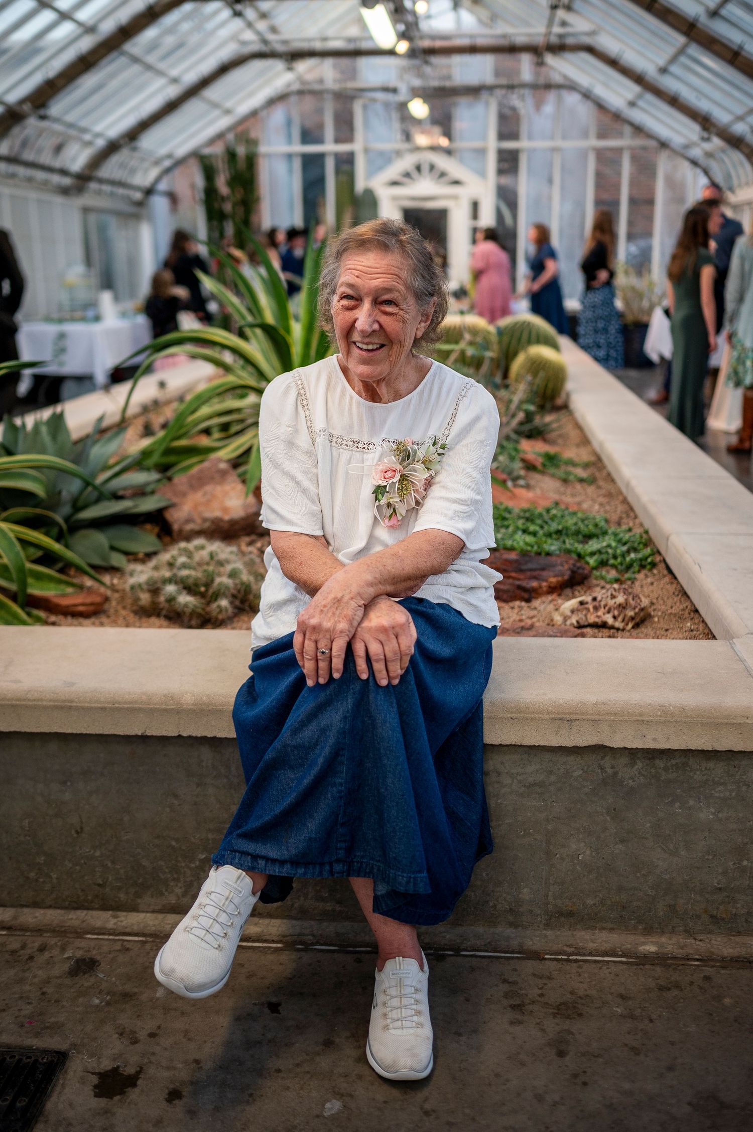 An elderly person in a white shirt and blue skirt sits in a greenhouse filled with plants.