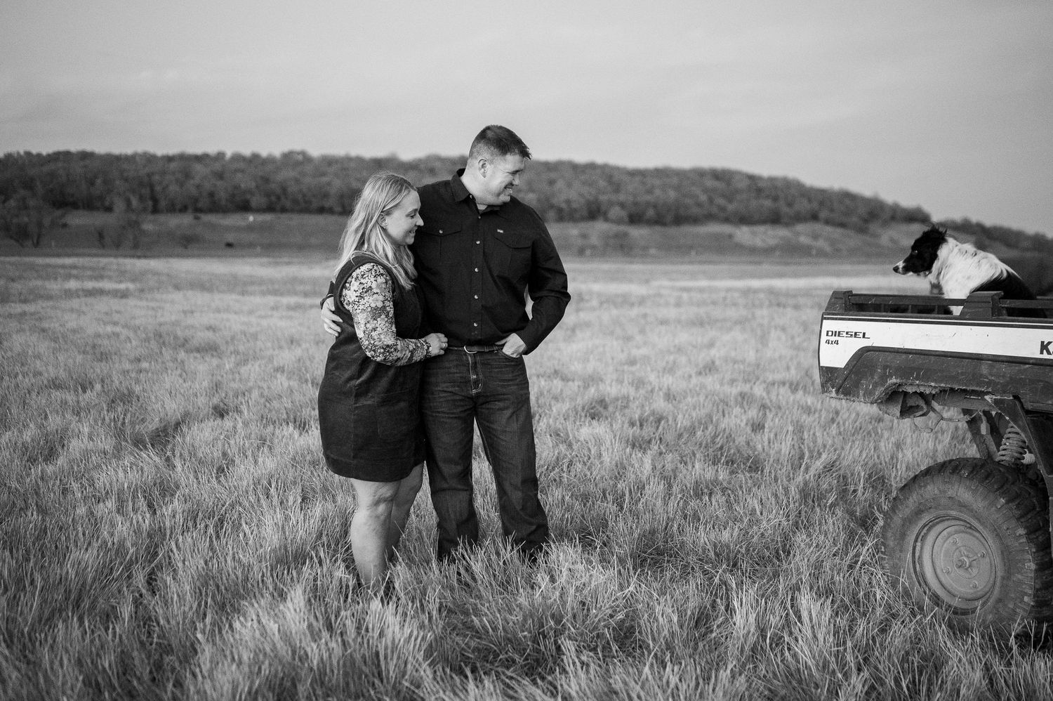 Black and white photo of a couple standing together in a field with a tractor nearby at dusk.