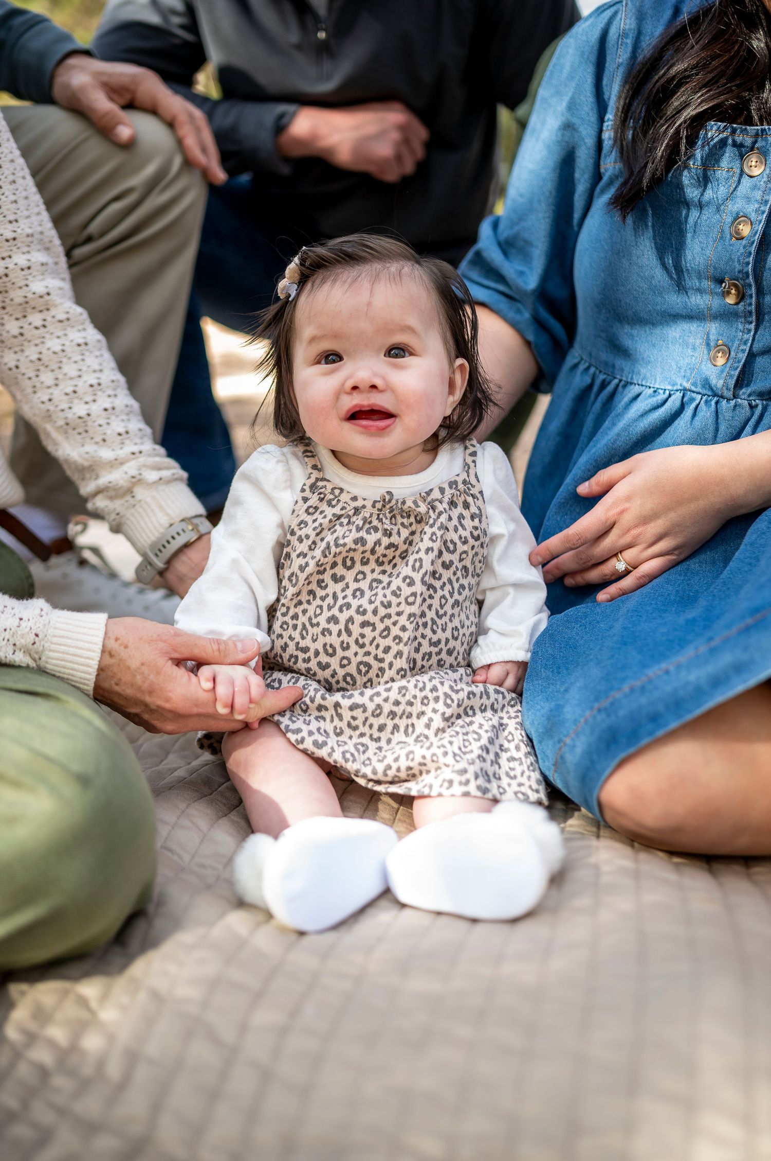 Smiling baby girl in leopard print dress sits on blanket surrounded by family members during outdoor photo session.