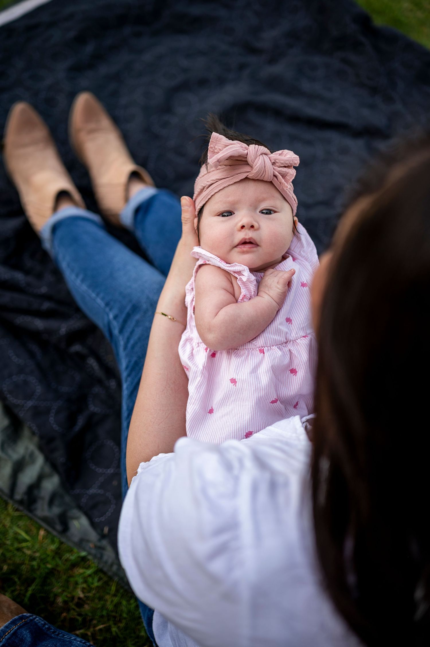 An infant wearing a pink bow headband and dress lies on a black blanket while being held by someone wearing jeans.