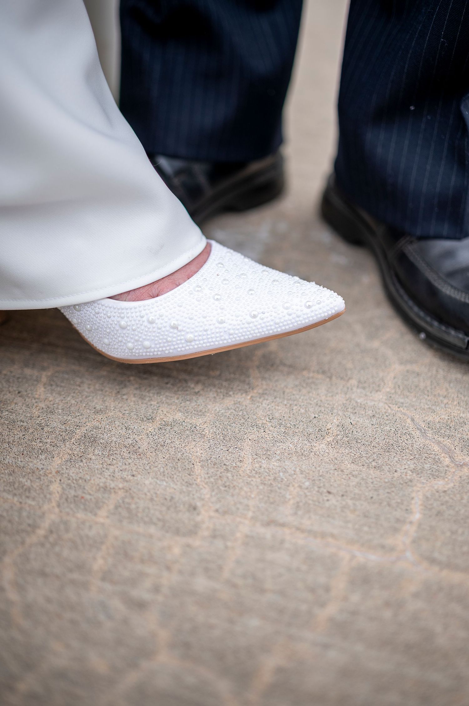 Close-up detail shot of wedding shoes, showing a white bridal flat next to formal black dress shoes.