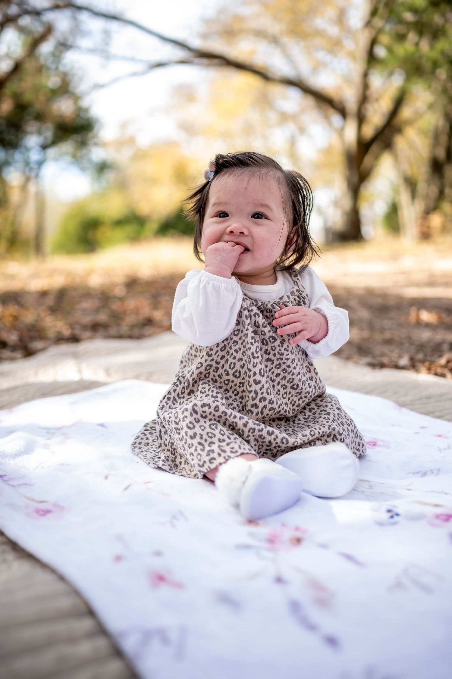 Adorable baby girl in leopard print dress sitting on white blanket outdoors, smiling with hand near mouth.