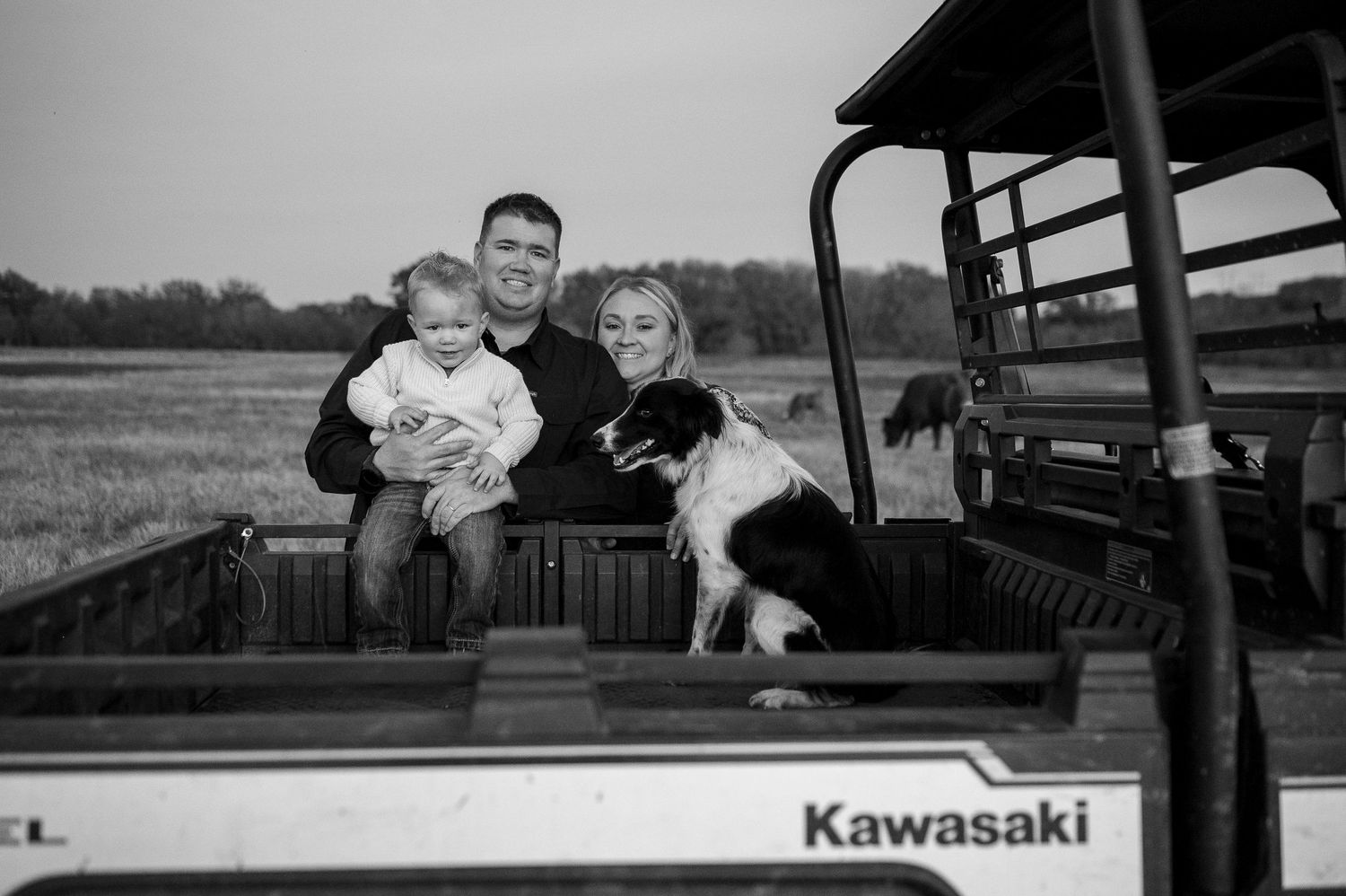 Family with baby and border collie dog posing on Kawasaki utility vehicle in farm field black white photo.