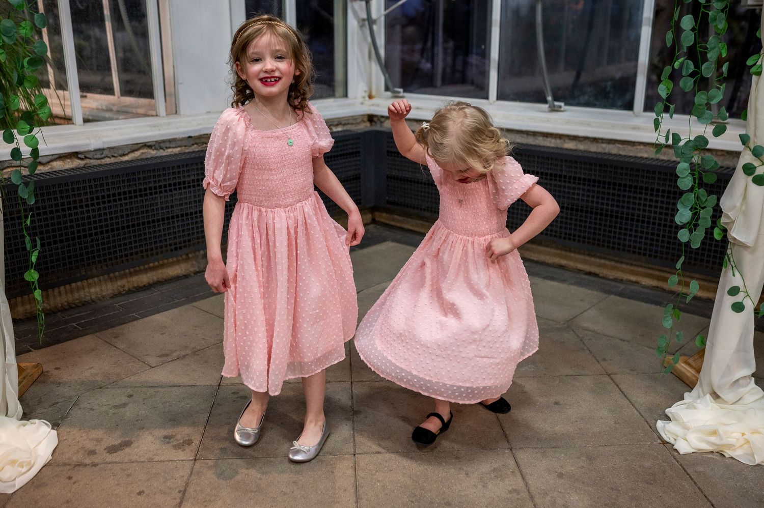 Two young girls in pink dresses twirling and dancing in a greenhouse setting.