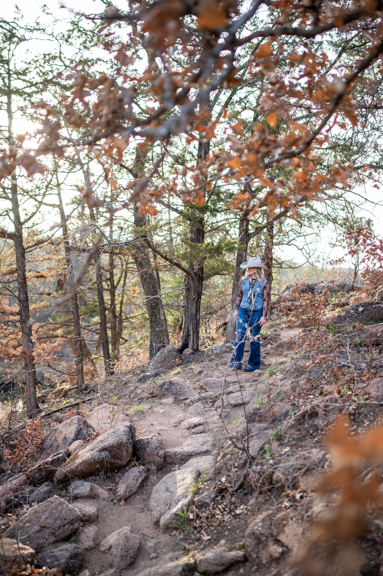 Child in hat and jeans hiking a rocky forest trail surrounded by autumn trees with orange leaves.