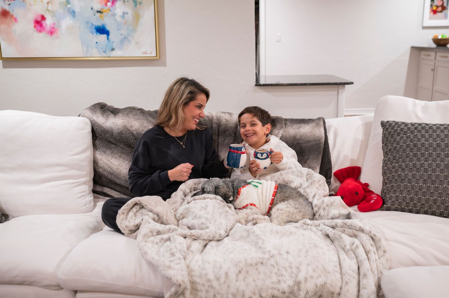 Two people snuggled under cozy blankets on a white couch sharing snacks.