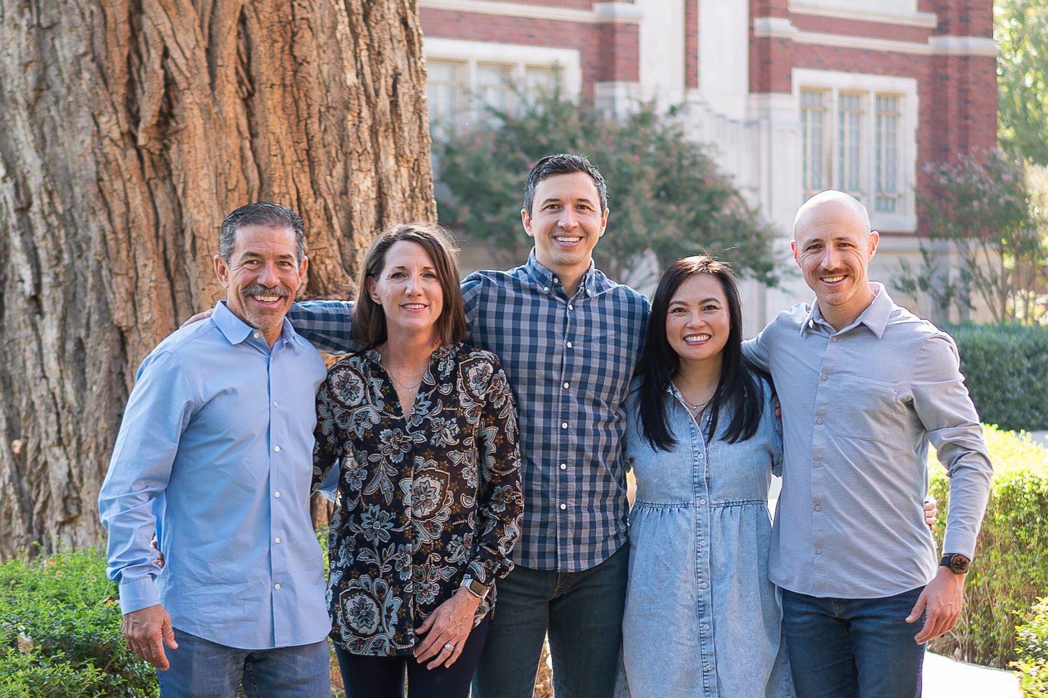 Group portrait in front of large tree trunk with collegiate brick building visible in background.