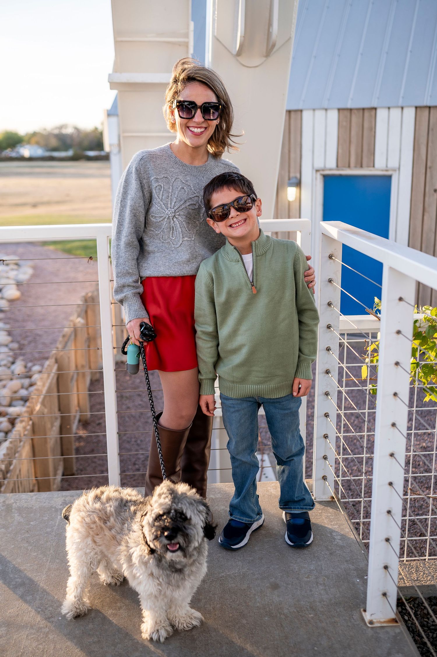 Woman and boy in sunglasses pose with fluffy dog on porch, woman wearing red skirt and gray sweater, boy in green pullover.