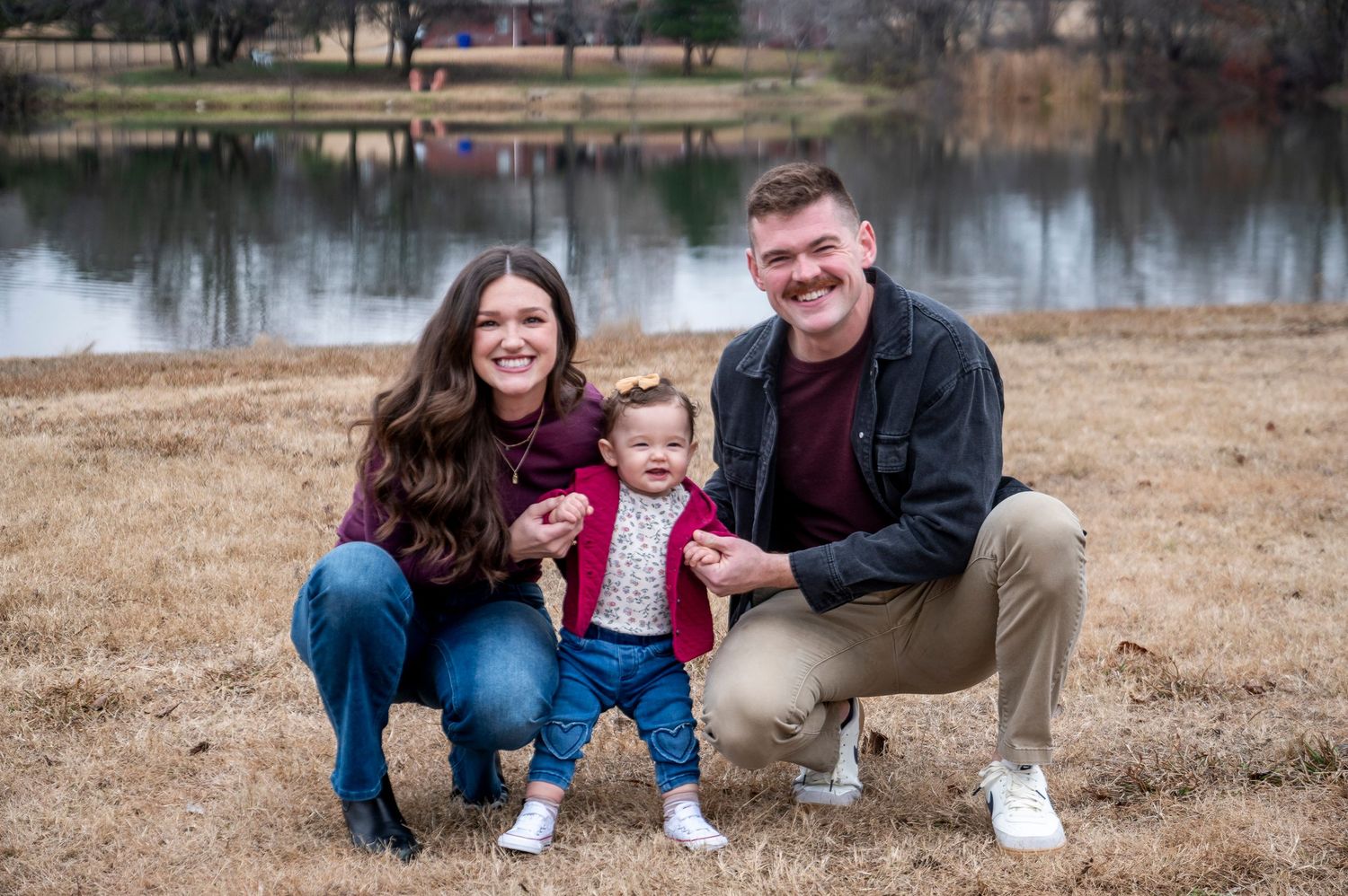 Family of three posing together by a calm lake during winter.
