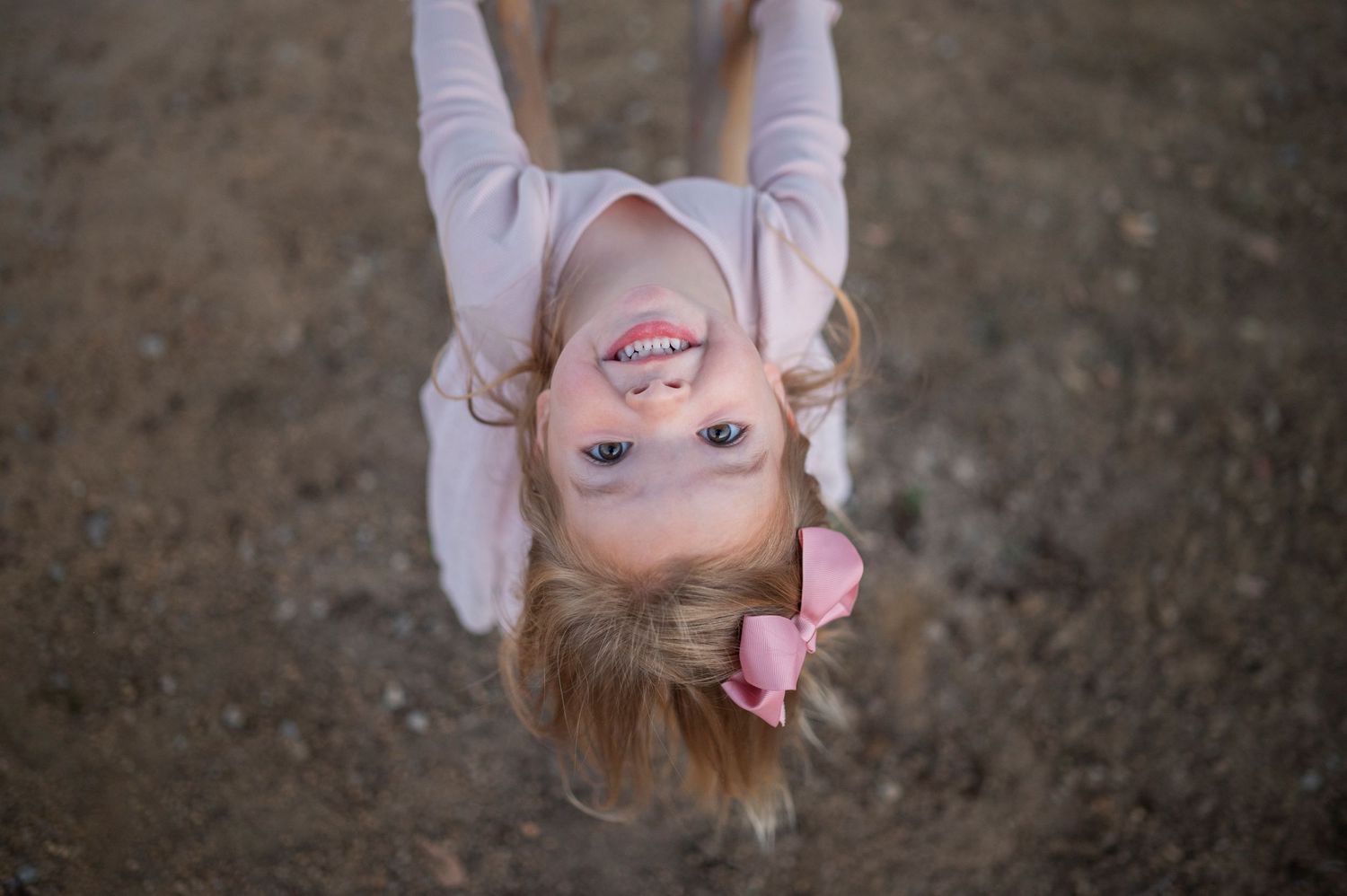 Playful upside-down portrait of a child with a bow hanging from playground equipment.