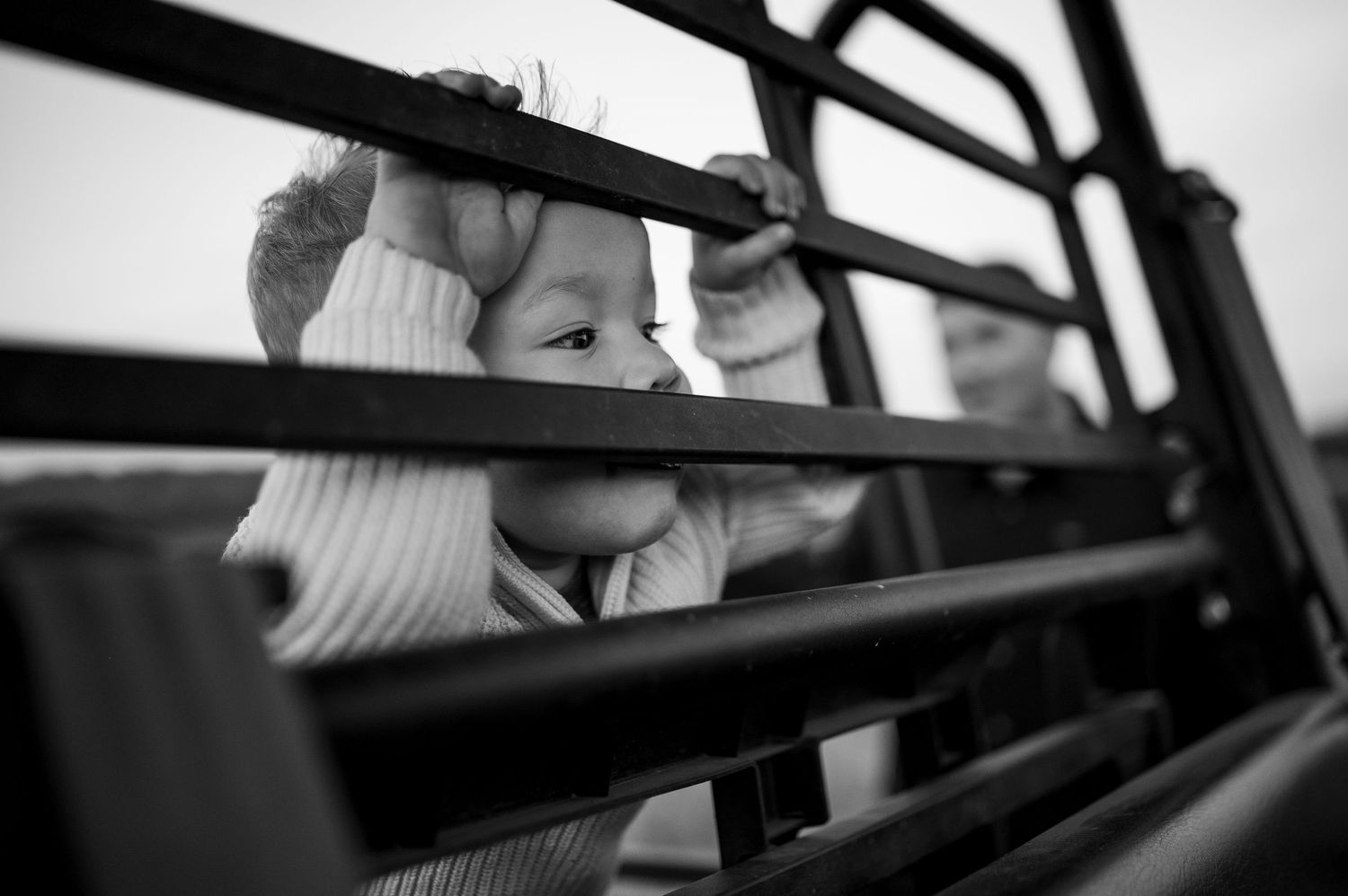 Black and white photo of a young child peering through metal bars of a vehicle, with another child visible behind.
