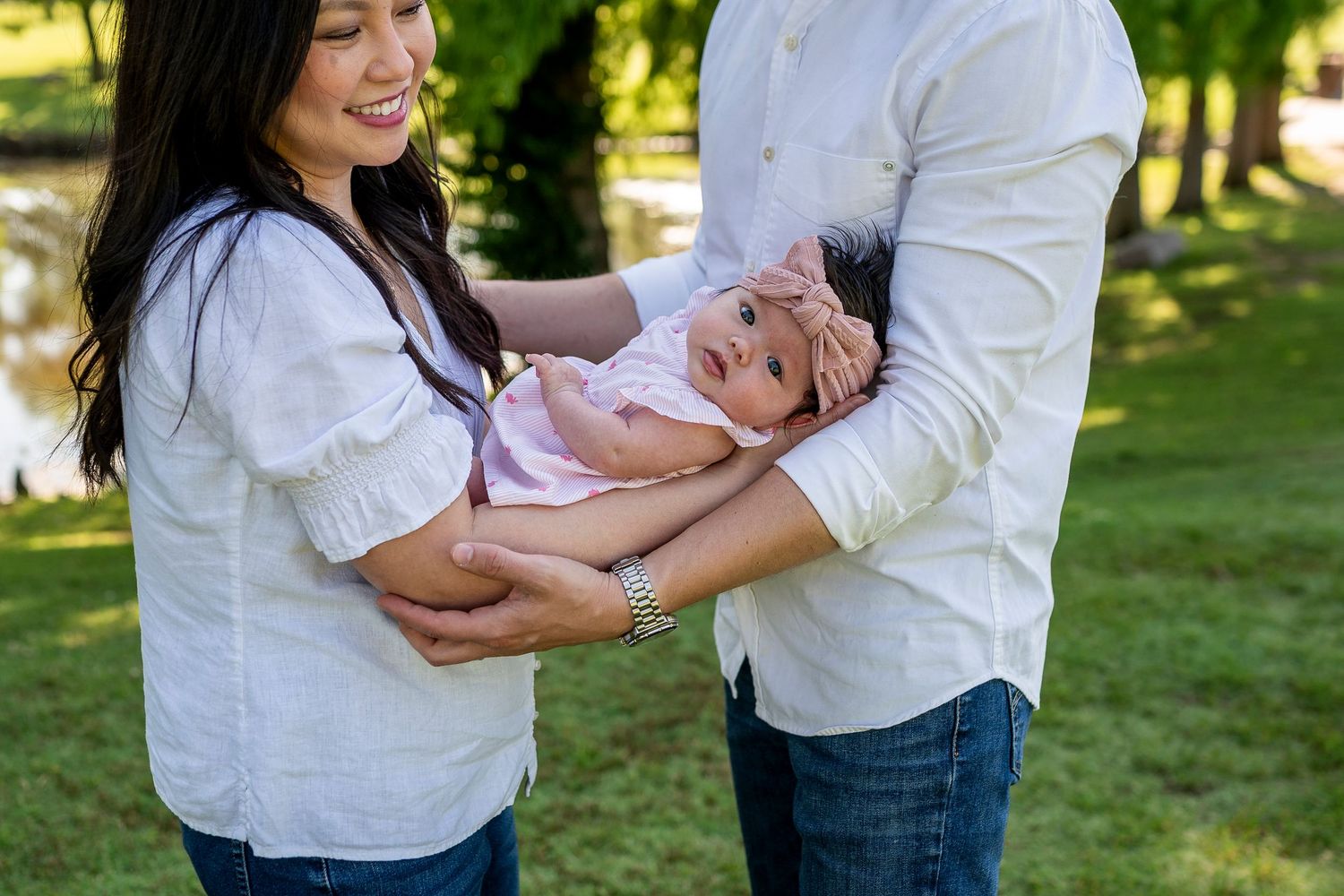 Parents in white shirts share joyful moment with baby in pink outfit outdoors.