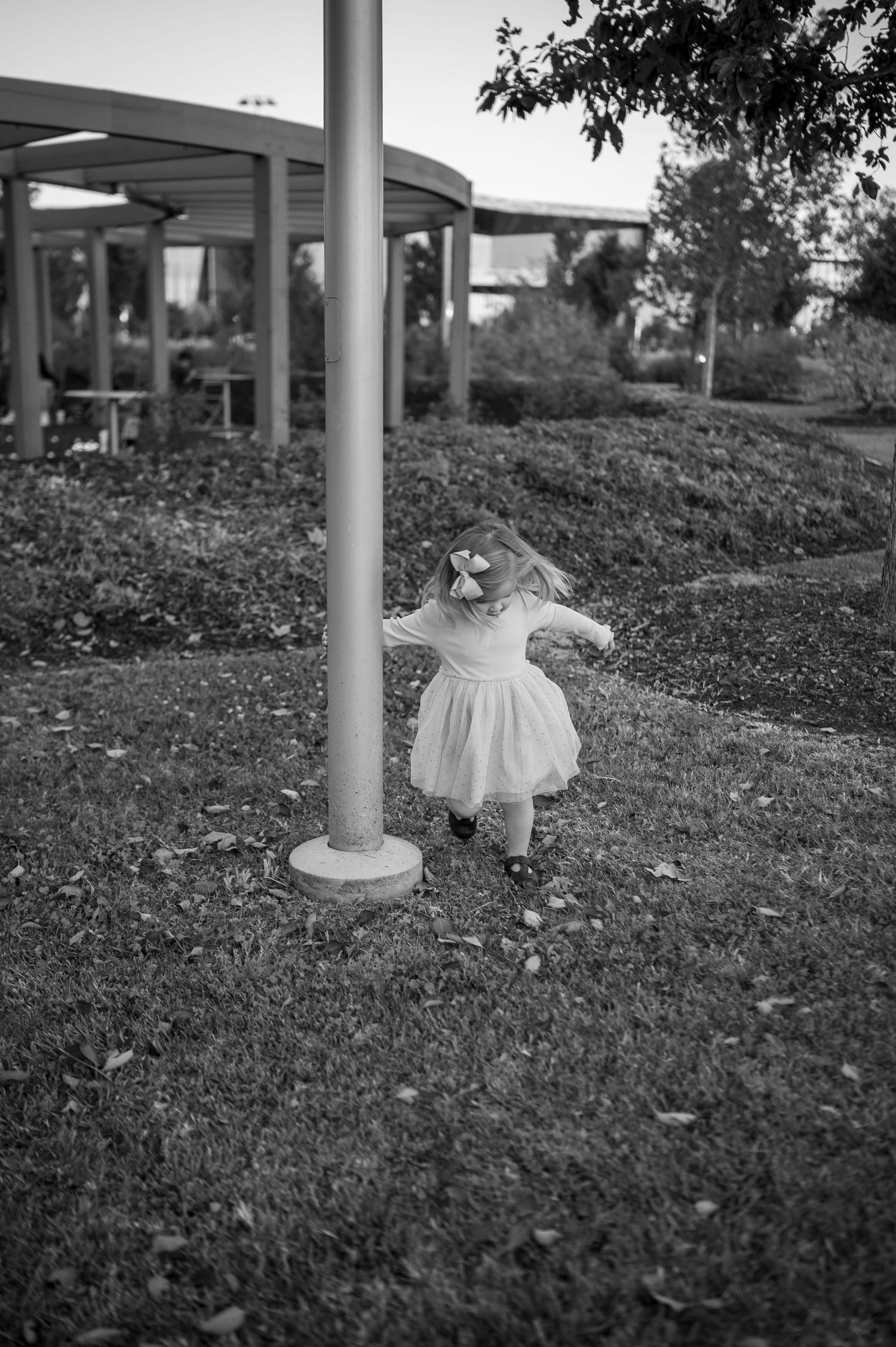Black and white photo of a small child in a dress playing near a metal pole outdoors.