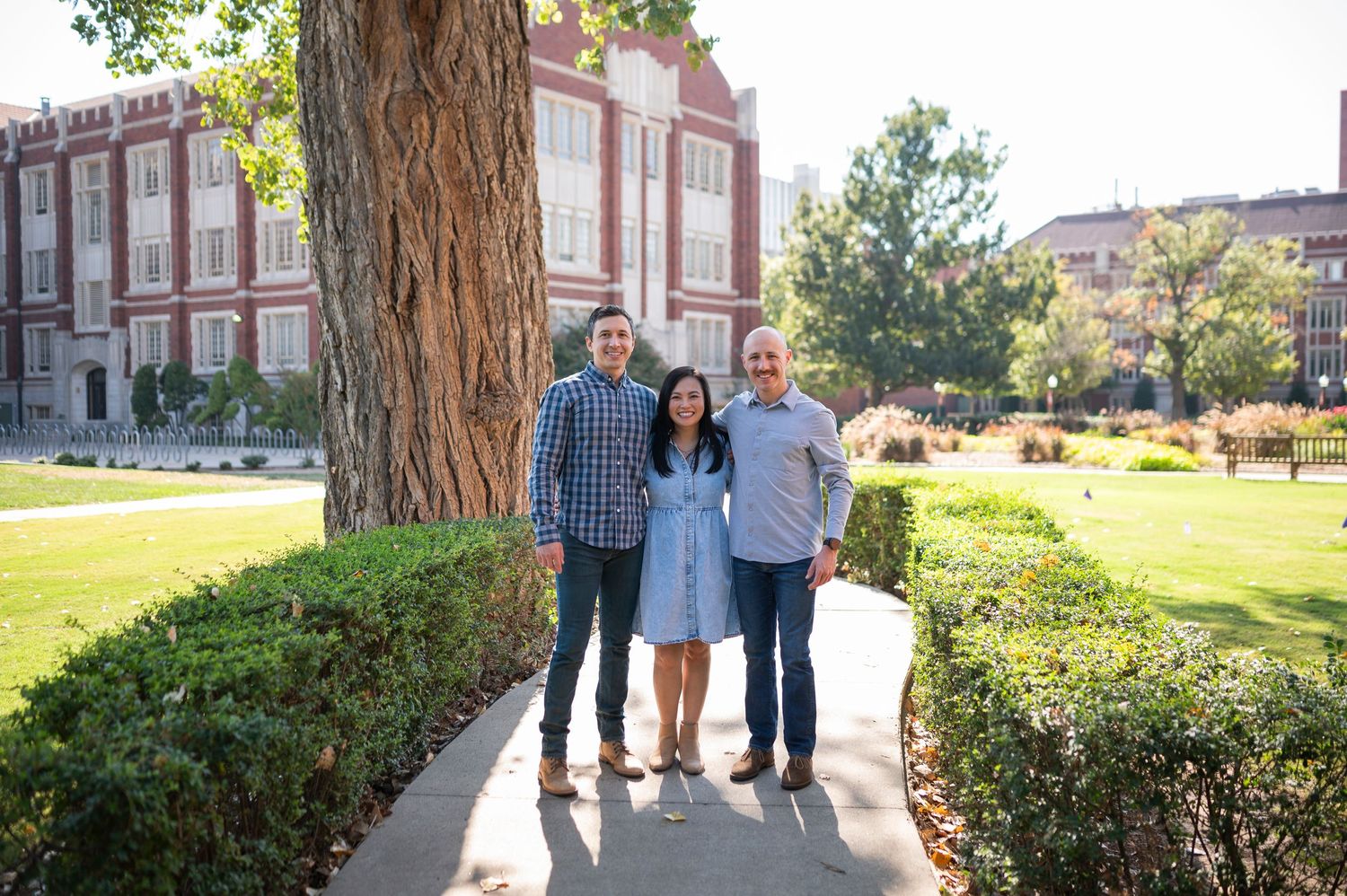 Three people standing on garden path with large tree trunk and collegiate building in background.