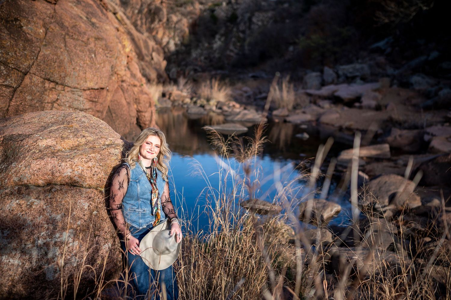 Woman with long blonde hair holding a hat stands by a rocky creek in a scenic outdoor landscape.