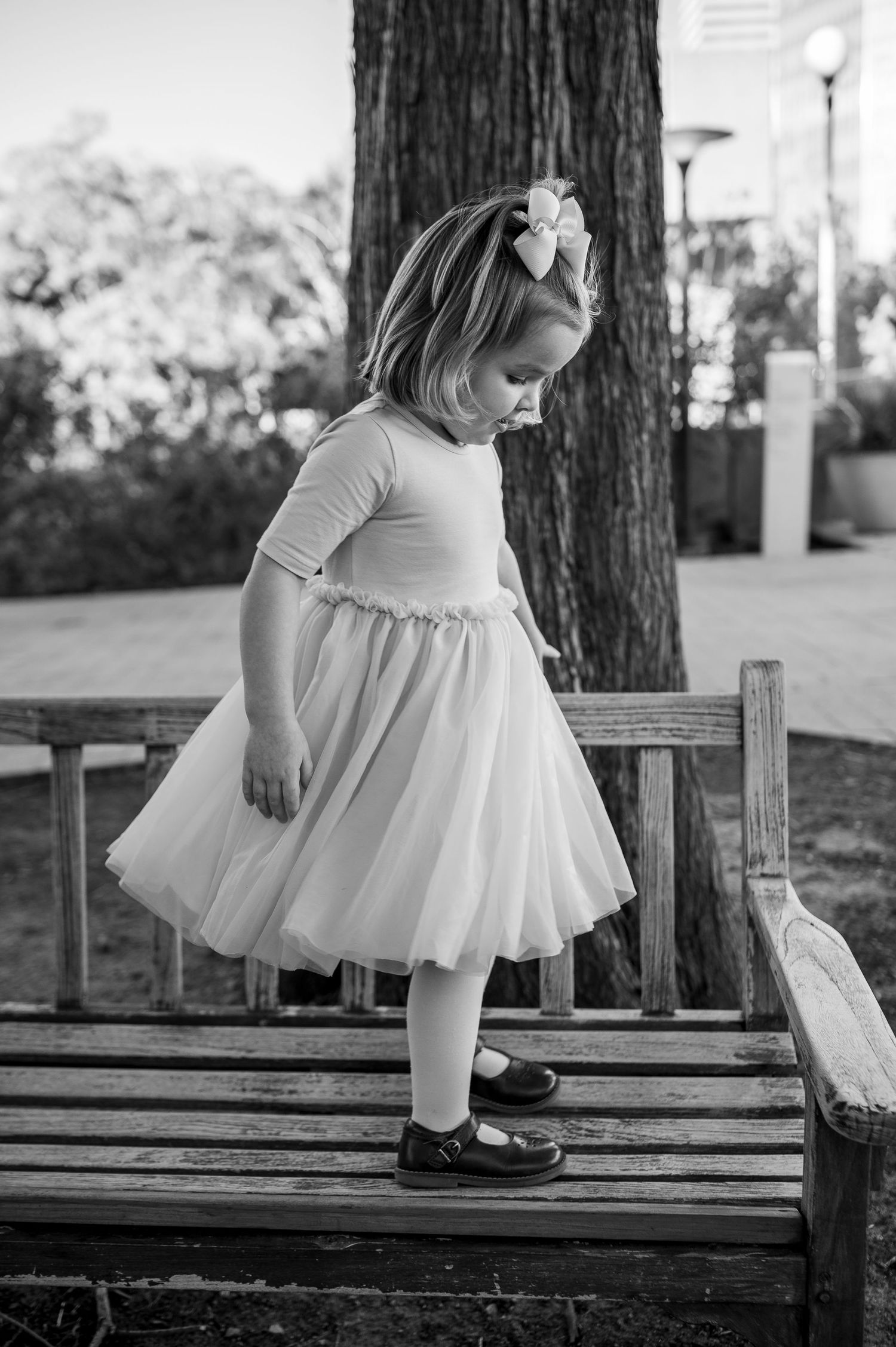 A young girl in a tutu dress and bow hairpiece stands on a wooden bench in a park, looking down.