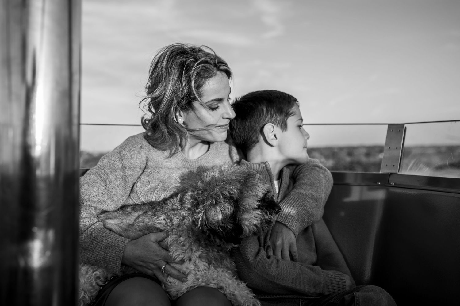 Black and white photo of a mother and child sharing a tender moment outdoors, with a small dog on her lap.