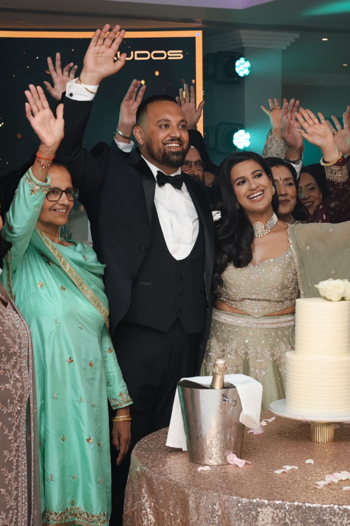 Wedding celebration with couple and guests cheering in front of a white cake at the reception.