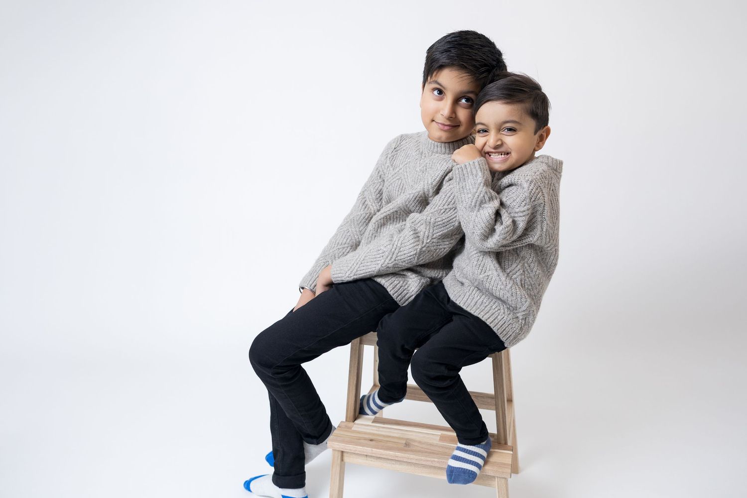 Two people in casual grey and blue outfits sit together on wooden furniture against a white studio backdrop.