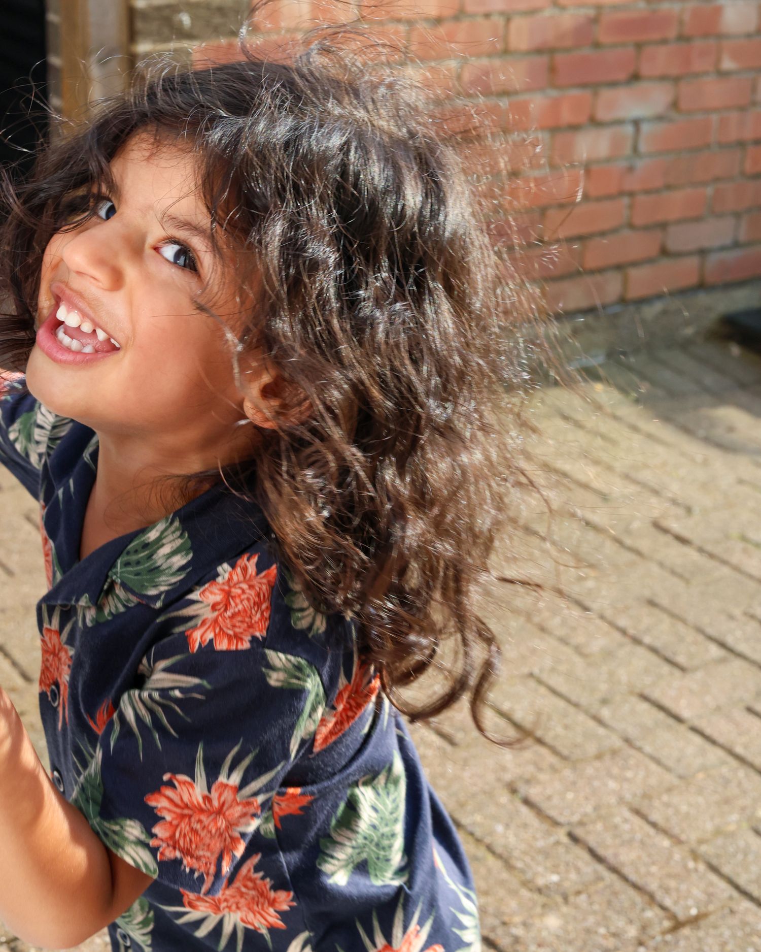 Child in floral print shirt laughing and playing outdoors against brick wall.