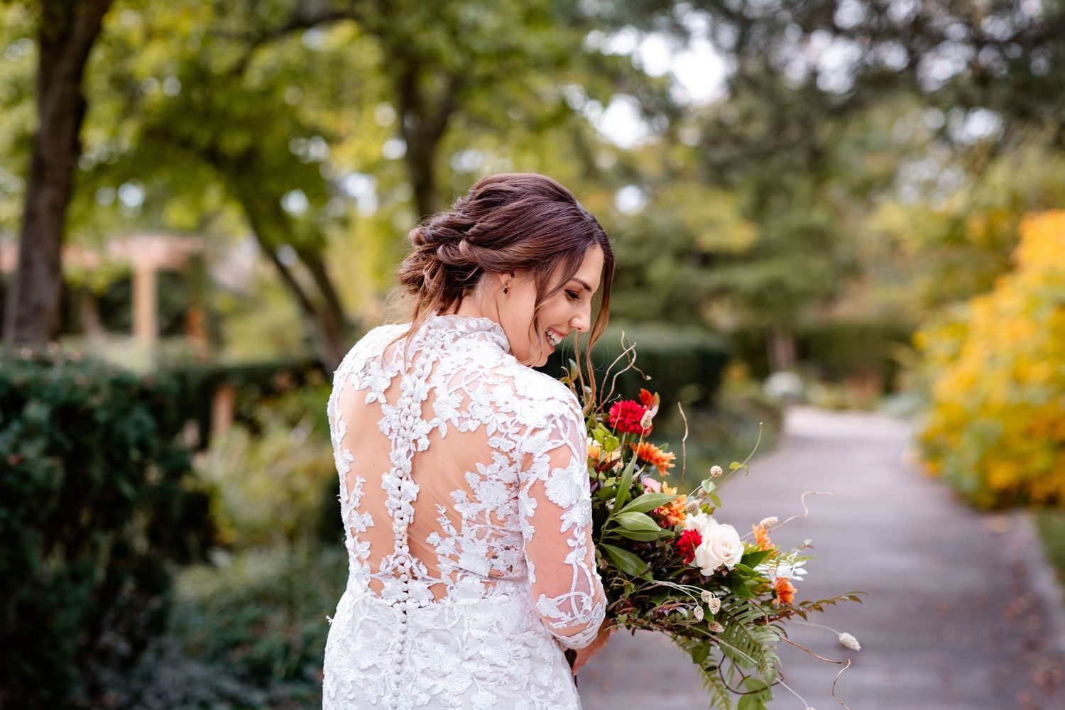 Bride in long-sleeve lace wedding gown holds vibrant floral bouquet on garden pathway.
