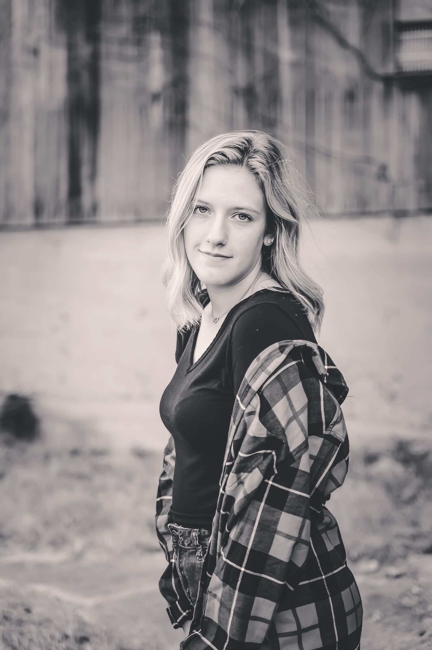 Black and white portrait of a blonde young woman in a plaid shirt, looking over her shoulder outdoors.