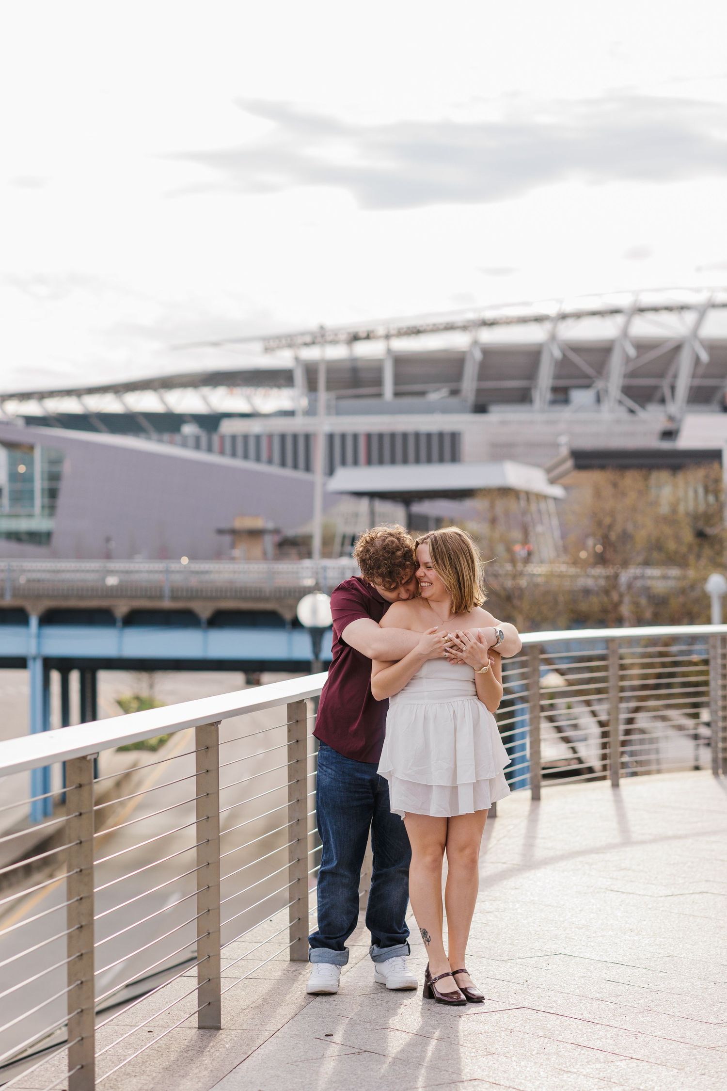 A couple shares a tender embrace on a waterfront boardwalk with Cincinnati Bengals stadium visible in the background.