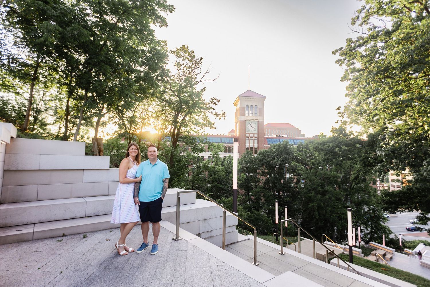 Couple posing on outdoor steps with a scenic view of trees and a historic building tower at sunset.