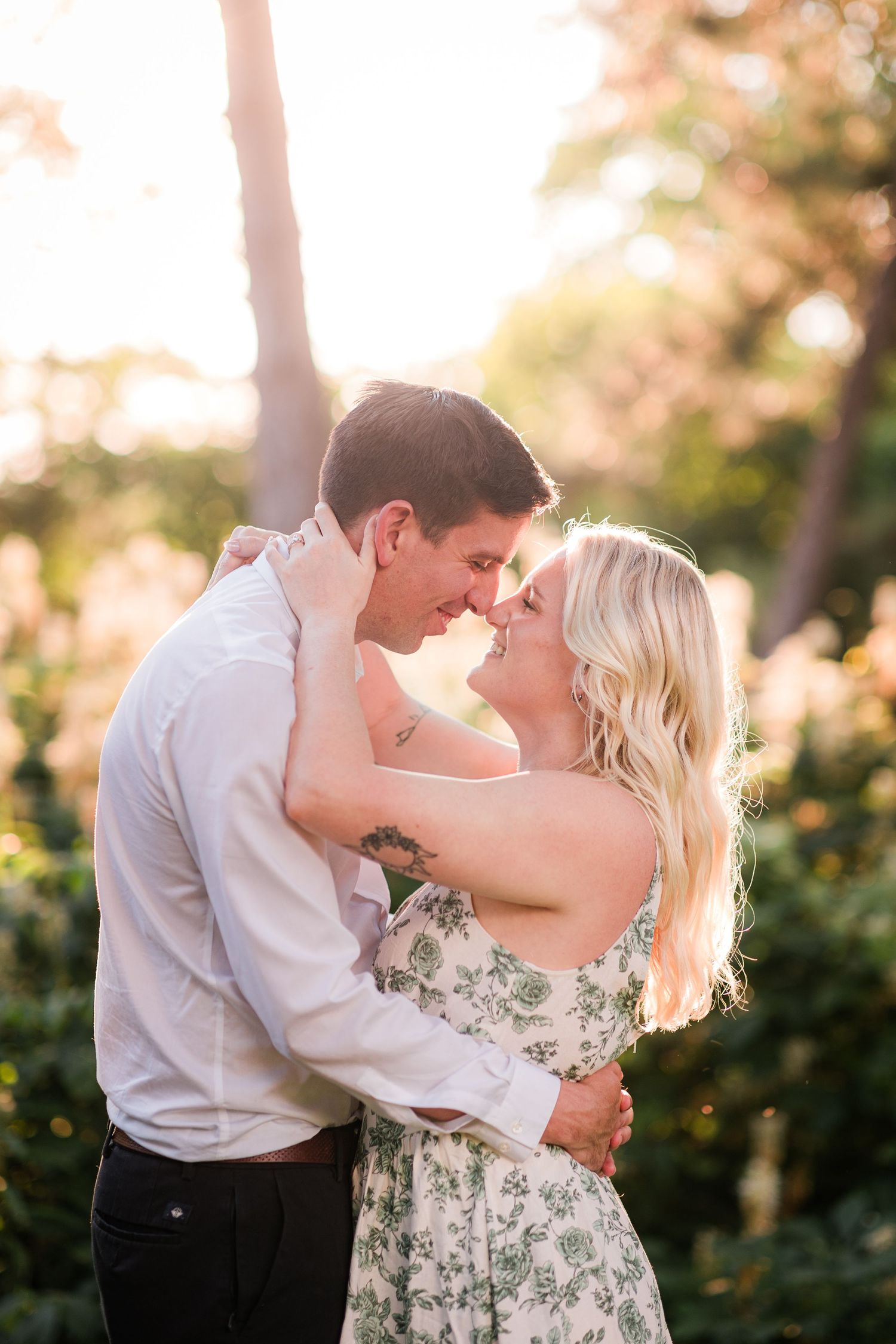 Couple sharing a romantic nose-to-nose moment outdoors with golden sunlight filtering through lush green trees.