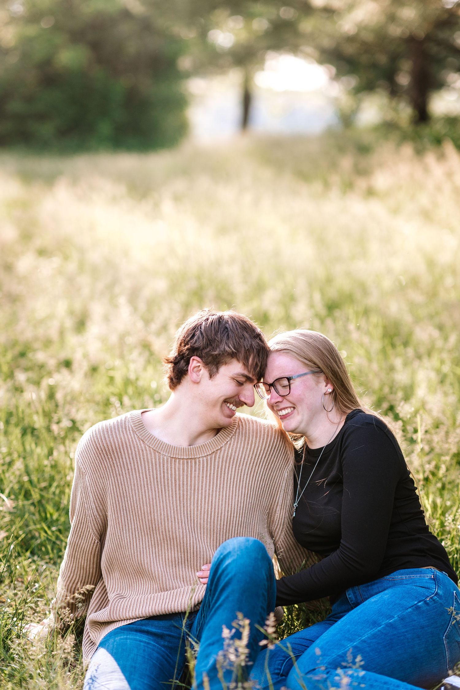 A happy young couple sits forehead to forehead laughing together in a golden sunlit grassy field.