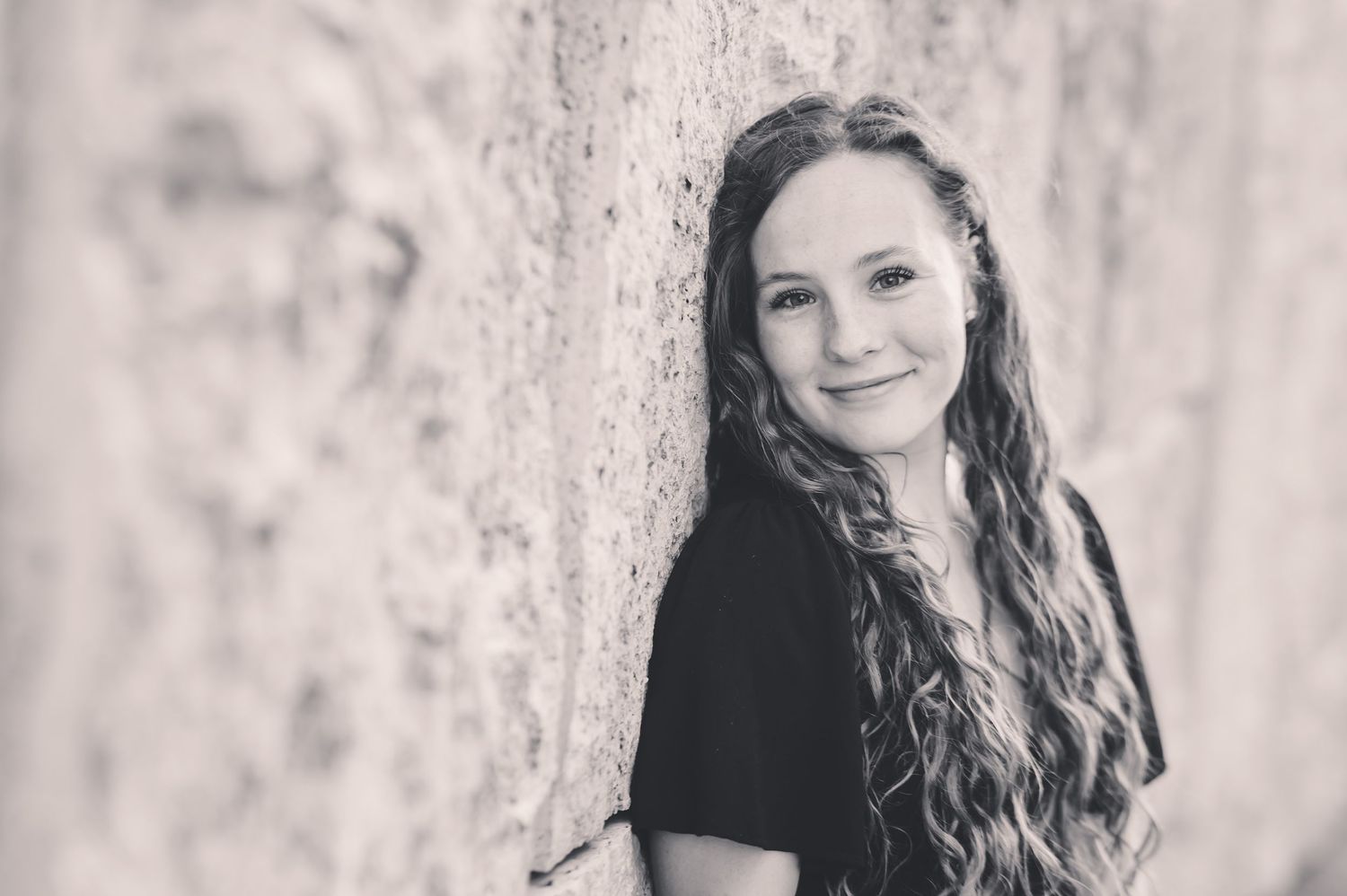 Black and white portrait of smiling young woman with long curly hair leaning on stone wall.