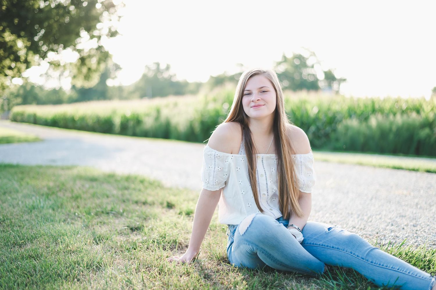 Teenage girl with long brown hair sits on green grass near a cornfield in a white off-shoulder top.