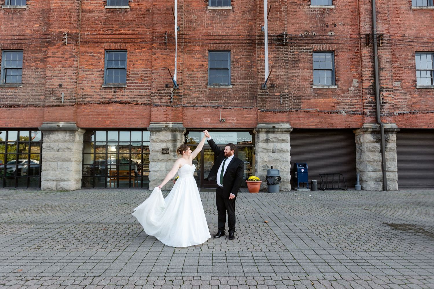 Bride and groom dance joyfully on cobblestone street in front of a historic red brick warehouse building.