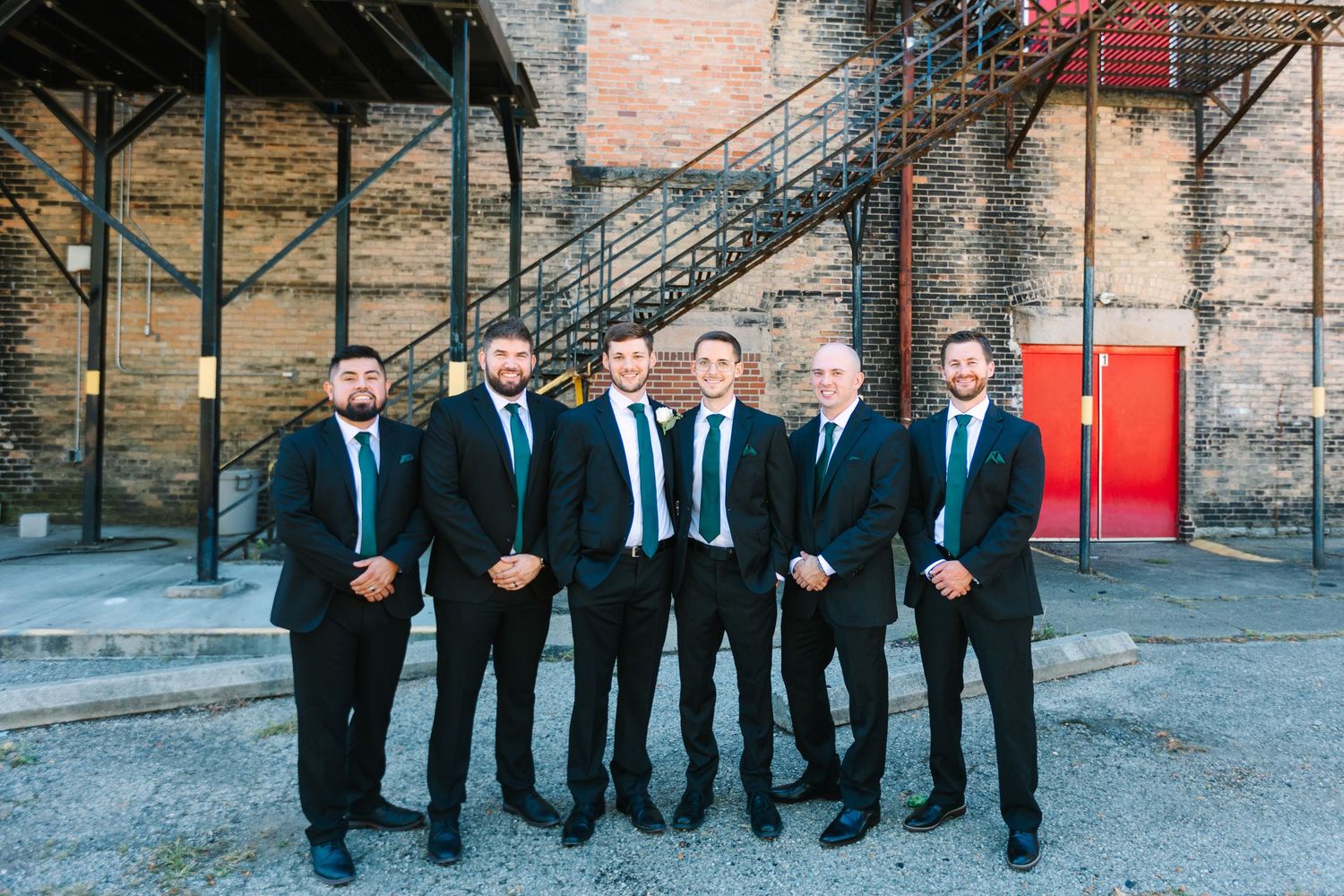 Groom and six groomsmen in black suits with teal ties posing outdoors near rustic brick building.