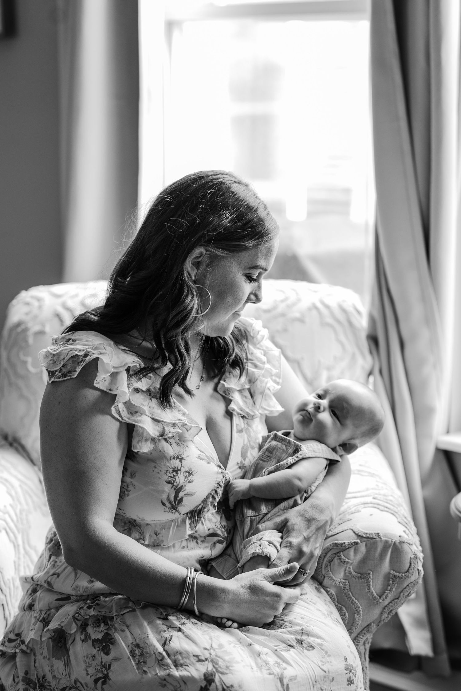 Black and white photo of a mother in floral dress tenderly holding her newborn baby by a window.