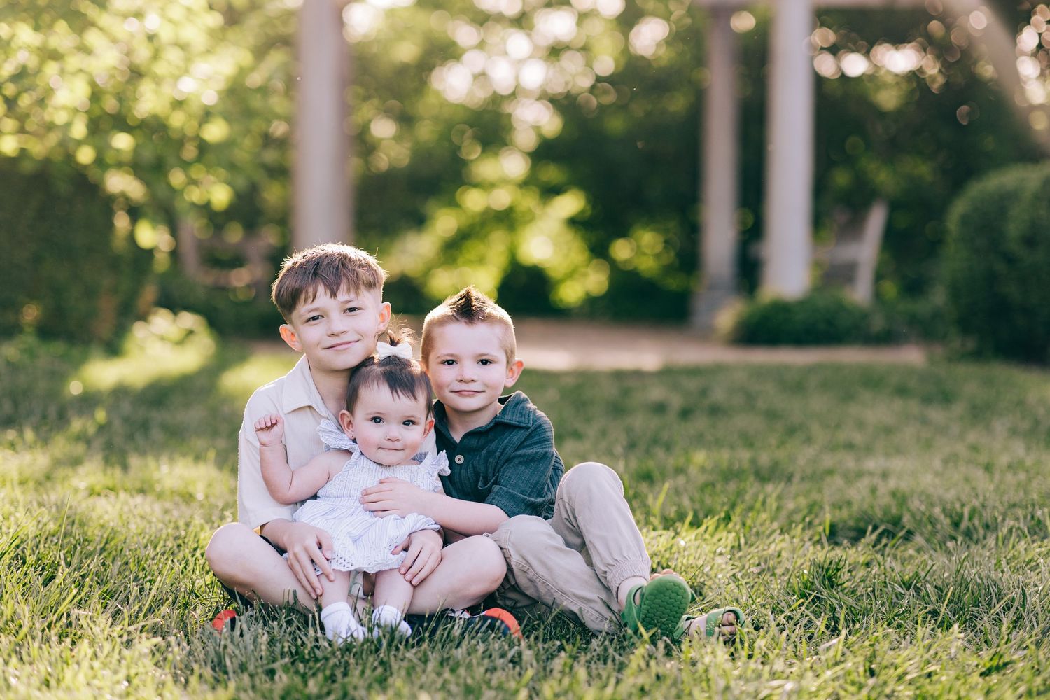 Two young brothers sitting on grass hugging their baby sister in a sunny garden.