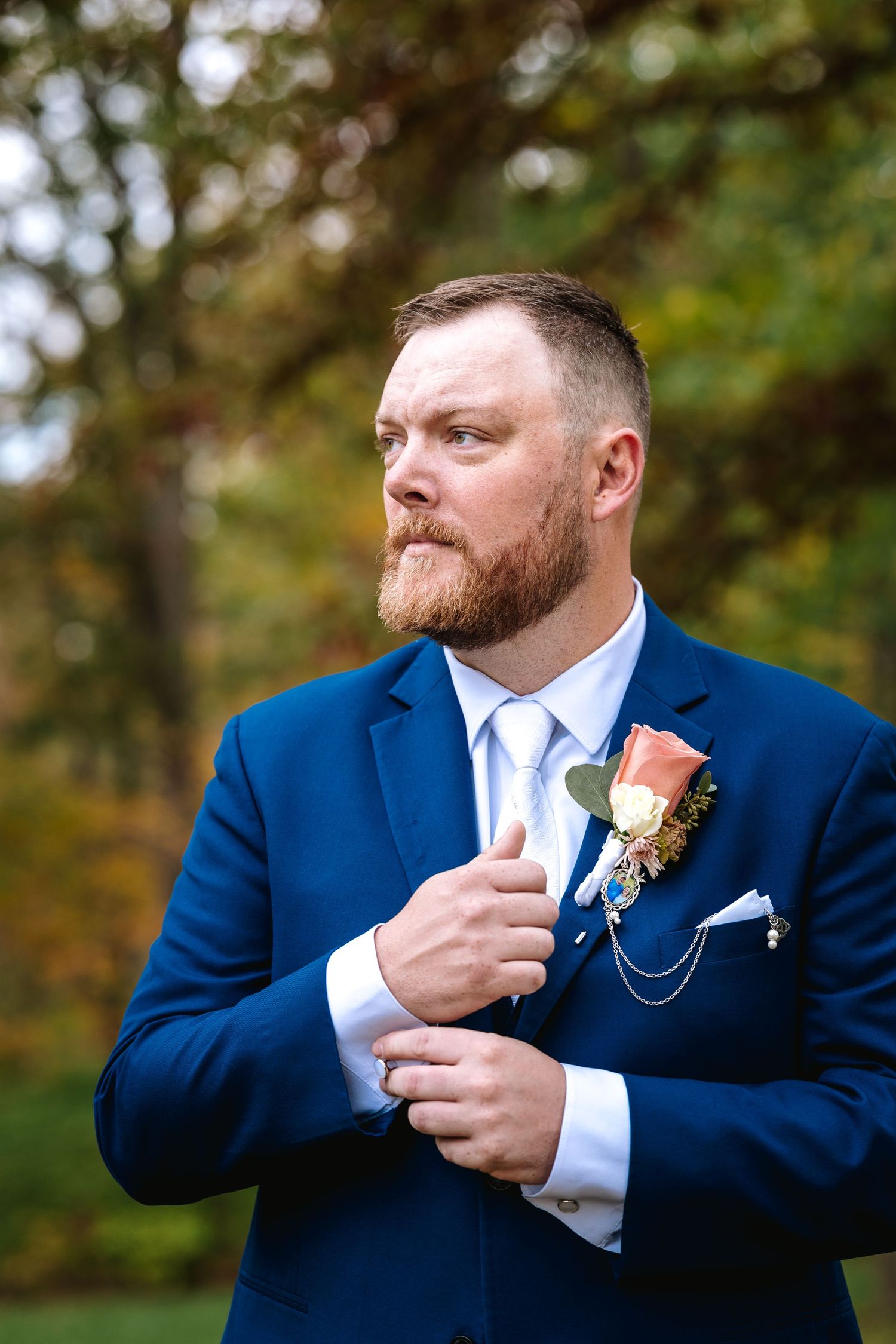 Bearded groom in navy blue suit adjusting cufflinks with floral boutonniere outdoors.