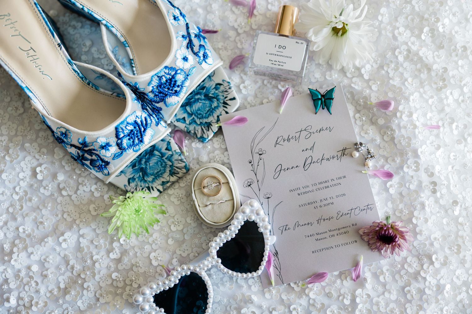 Wedding flat lay featuring blue floral heels, invitation suite, rings, and bridal accessories.