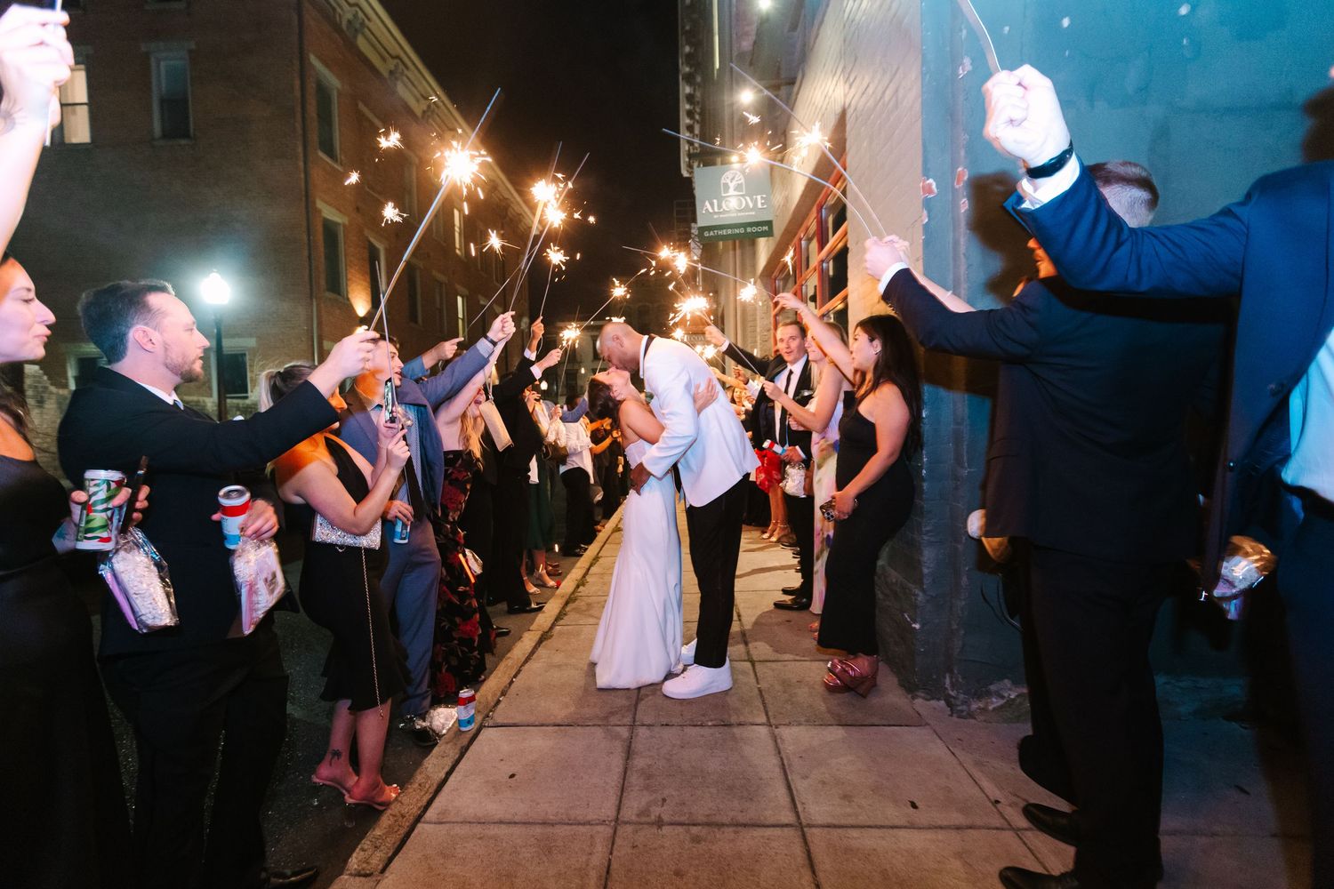 Bride and groom kissing during a sparkler exit surrounded by cheering guests at night.
