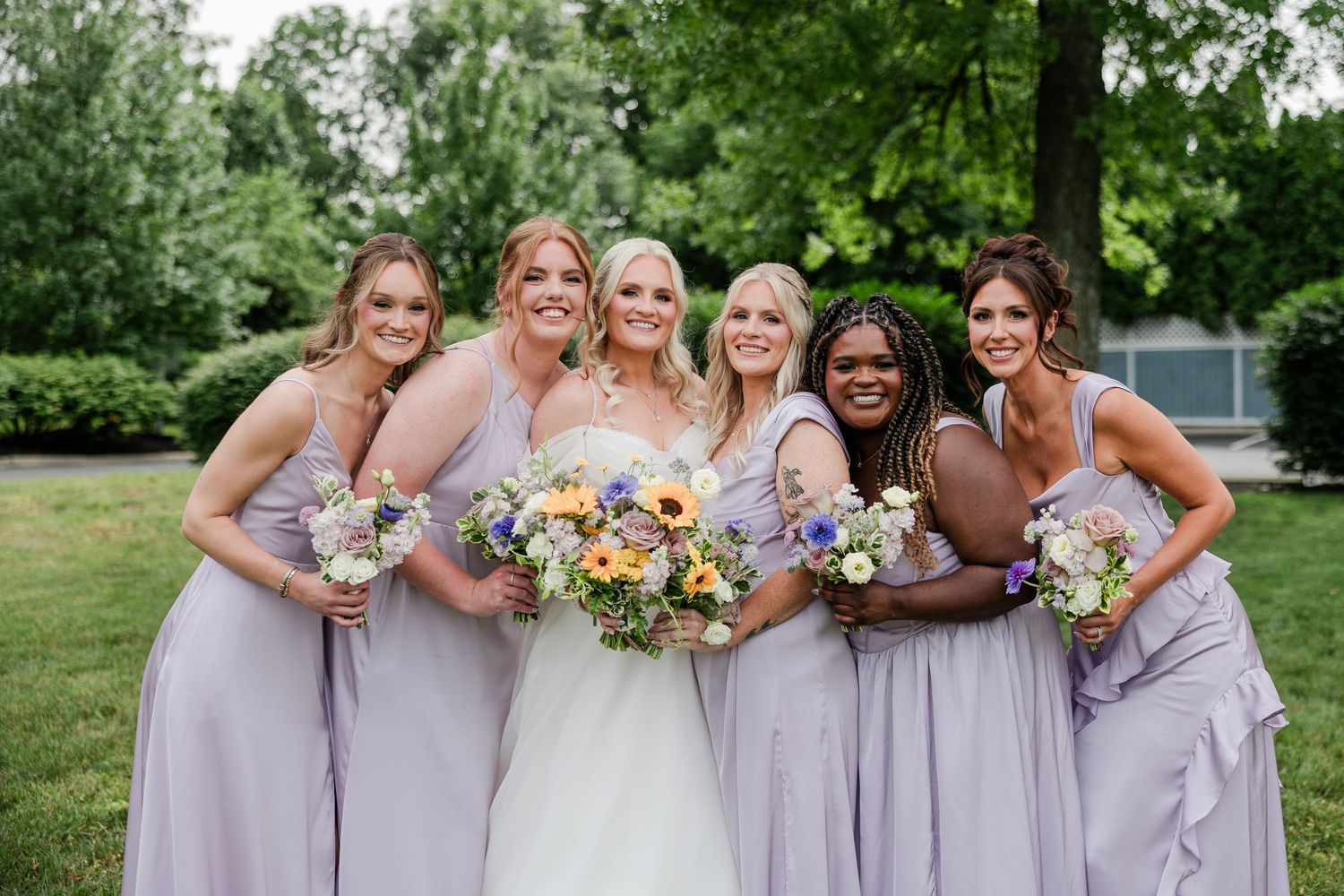 Bride posing with four bridesmaids in lavender dresses holding colorful floral bouquets outdoors.