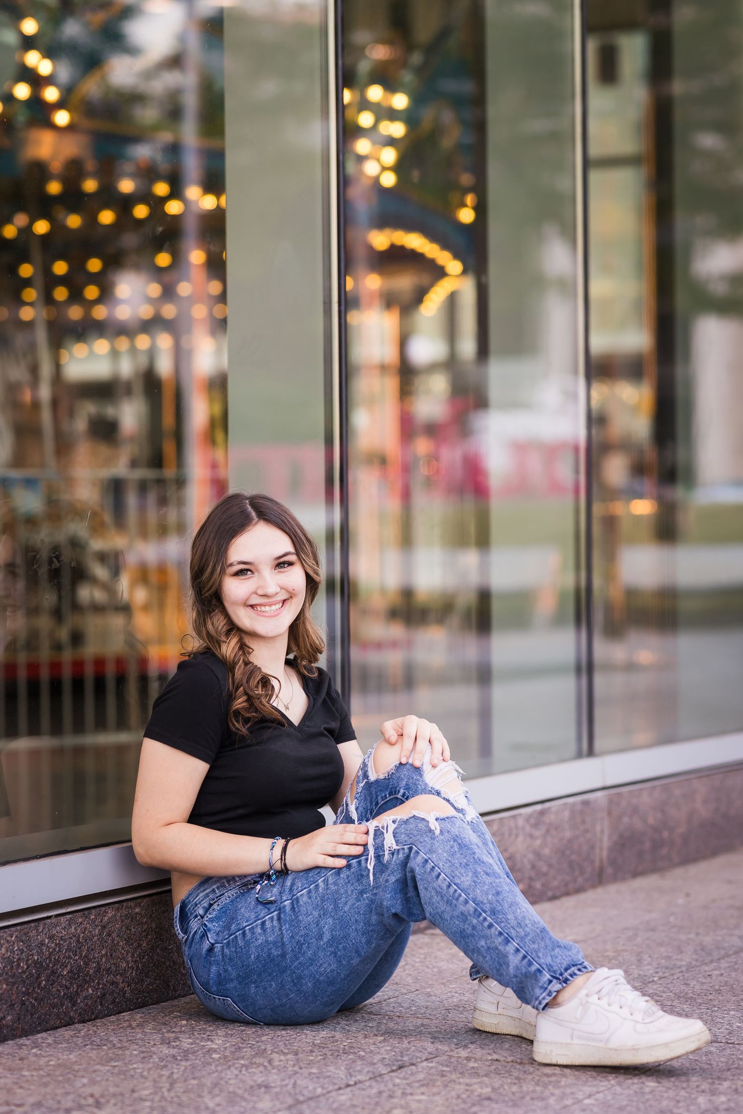 Smiling teenage girl in black top and ripped jeans sitting against a storefront window at dusk.